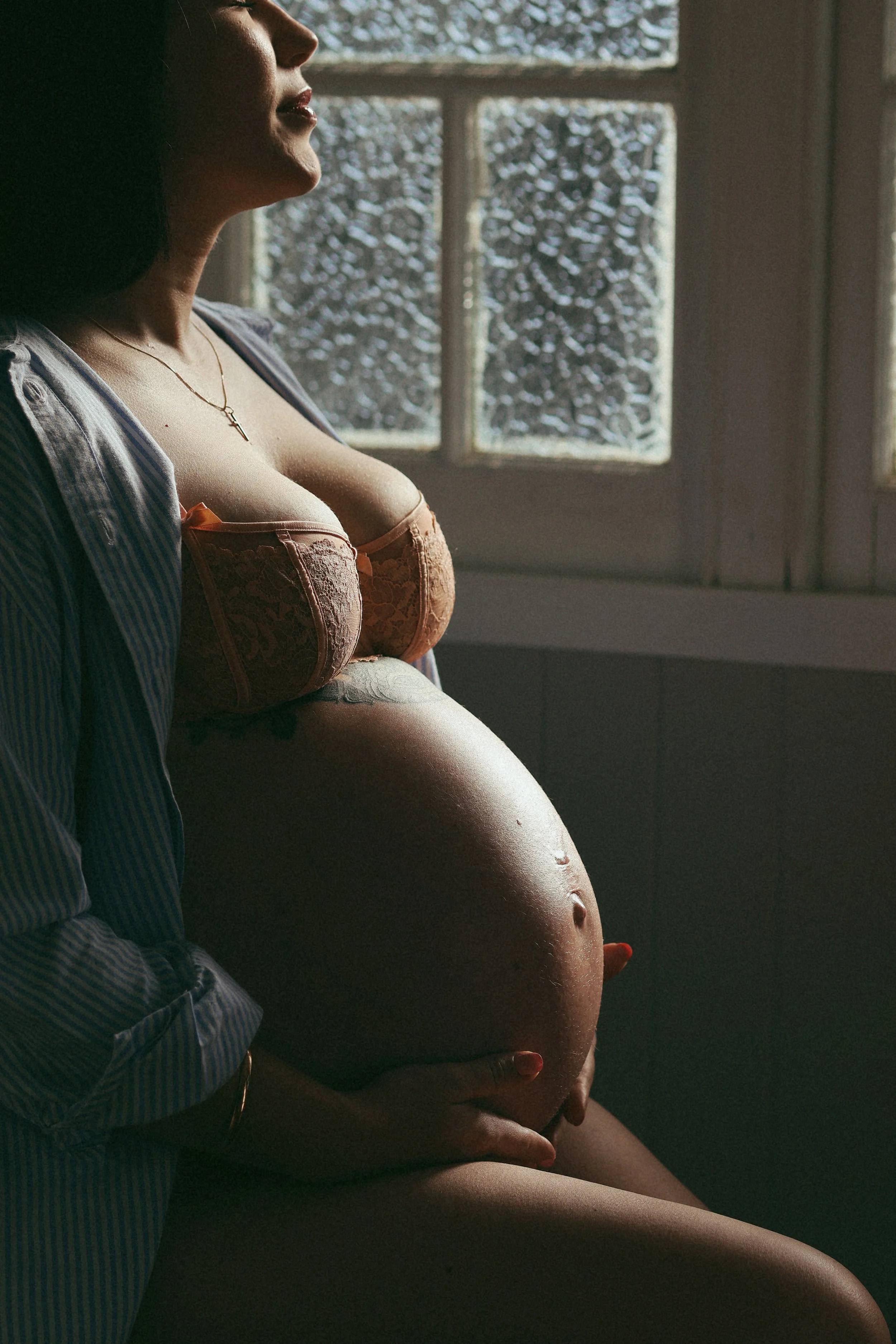A pregnant woman with a tattoo on her belly, wearing a peach-colored lace lingerie, sitting near a window with snowy scene outside, with a person standing beside her in a striped shirt and holding her belly.