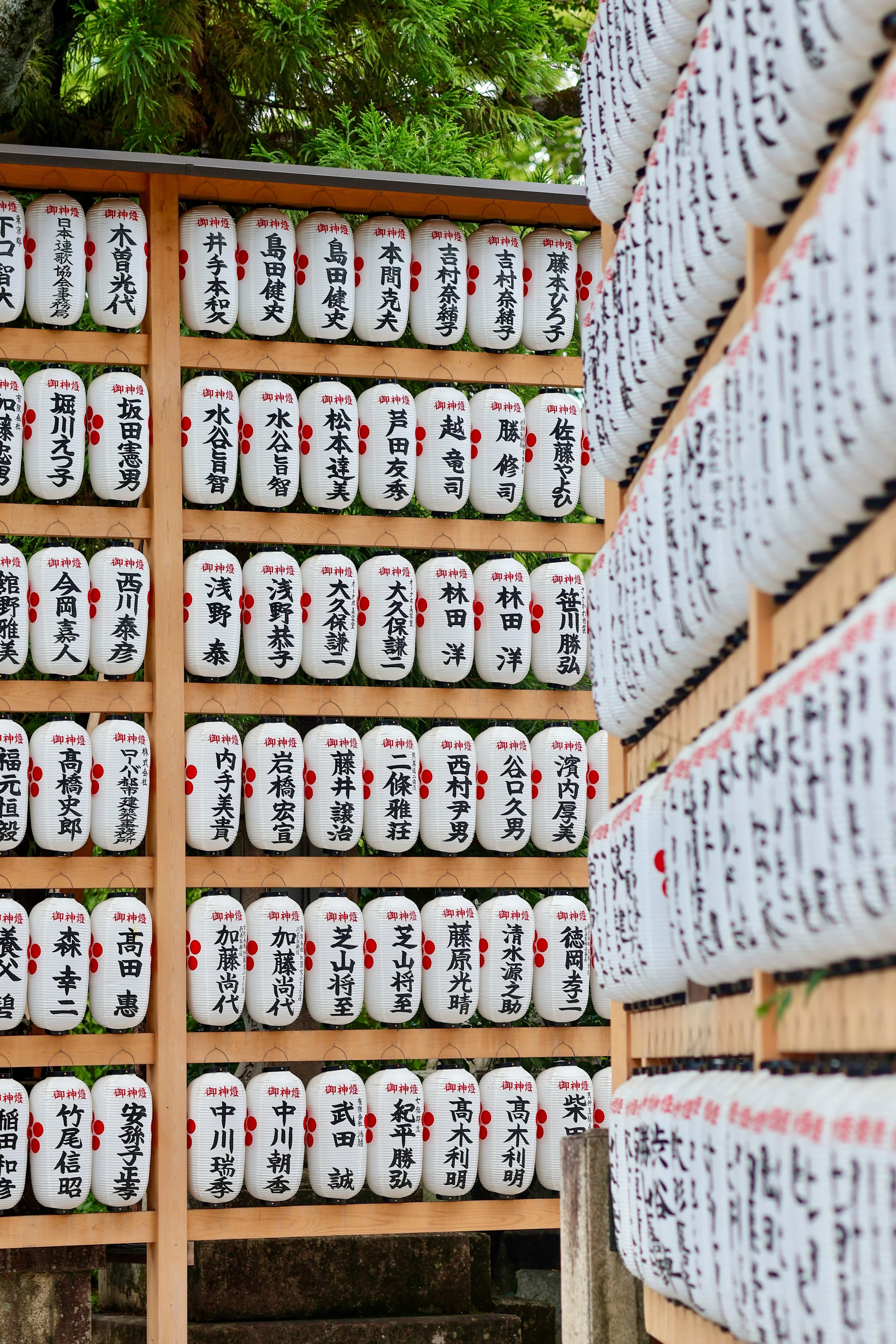 A display of traditional Japanese paper lanterns with black Japanese characters and red markings, arranged on a wooden frame, with green trees in the background.