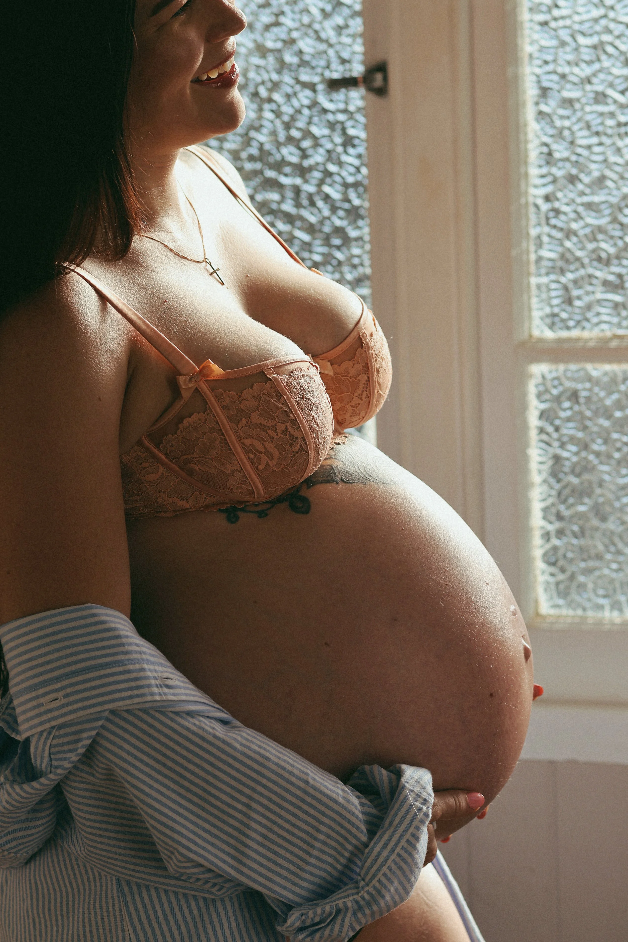 A close-up side view of a smiling woman in lingerie, with her pregnant belly being supported by a person wearing a striped shirt, near a window with patterned glass.