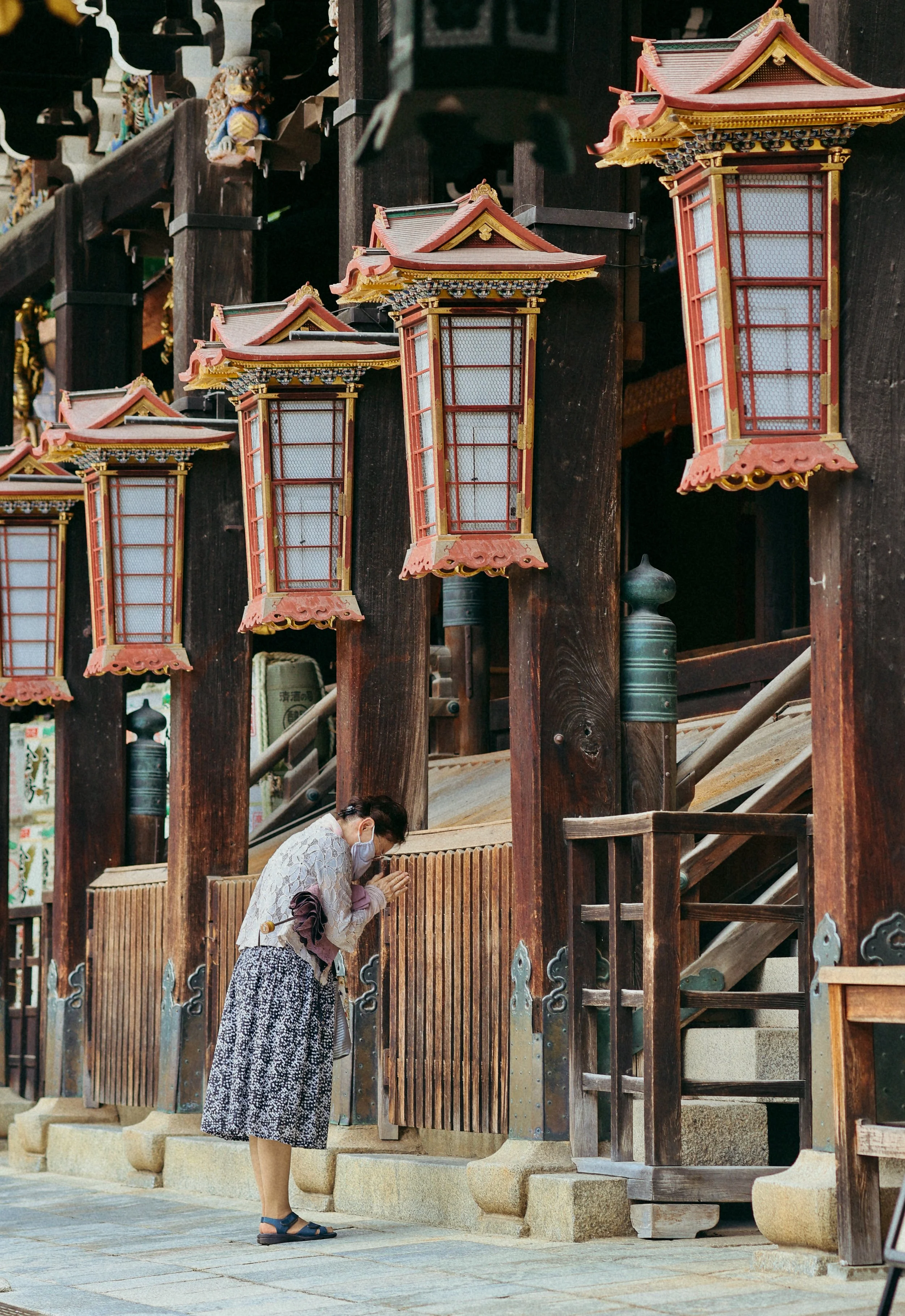 A woman praying at a traditional Japanese shrine, standing on stone pavement, with wooden structures and lanterns in the background.