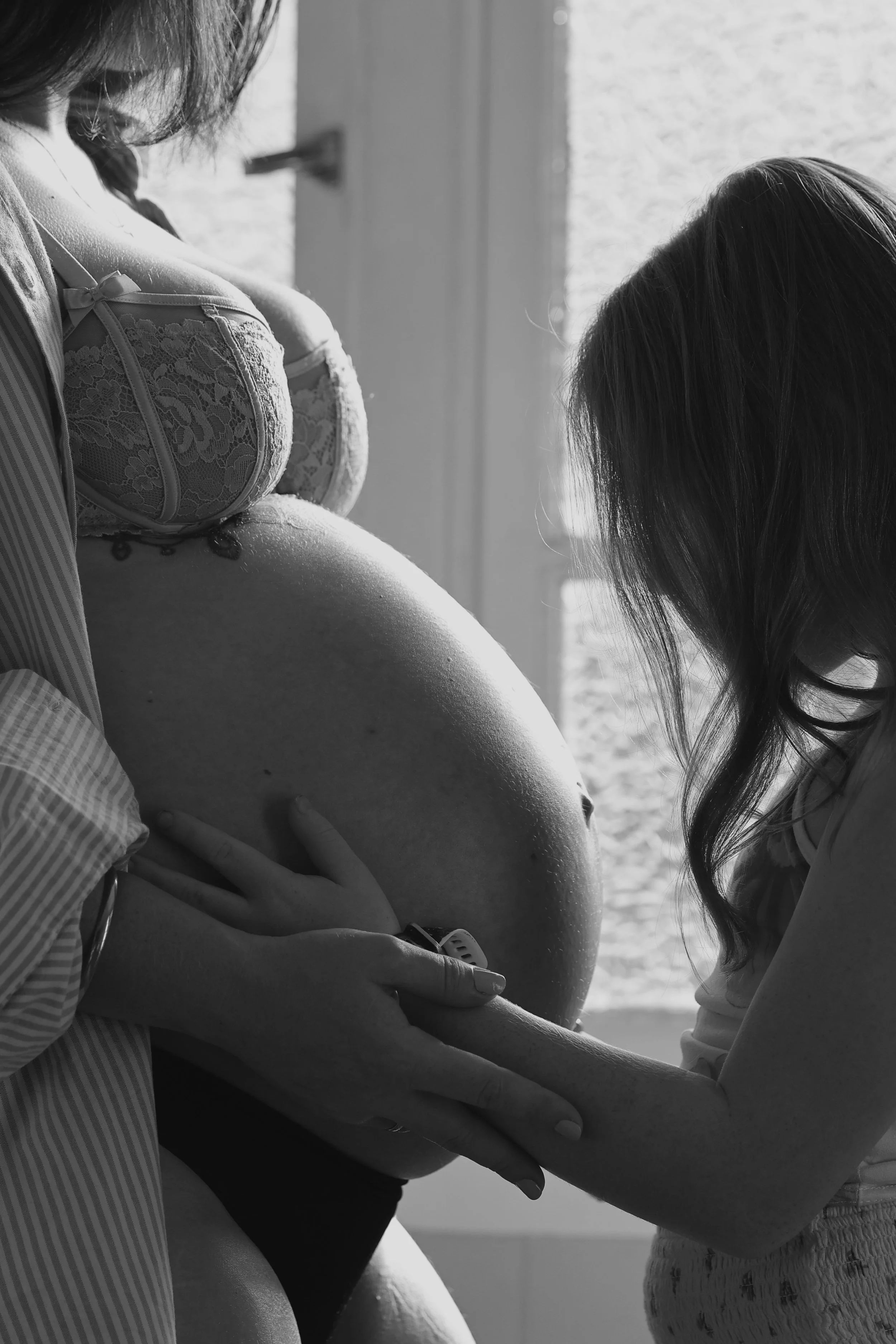 Close-up of a pregnant woman in lace lingerie with a woman touching her belly, in a bright room with sunlight and window blinds in the background.