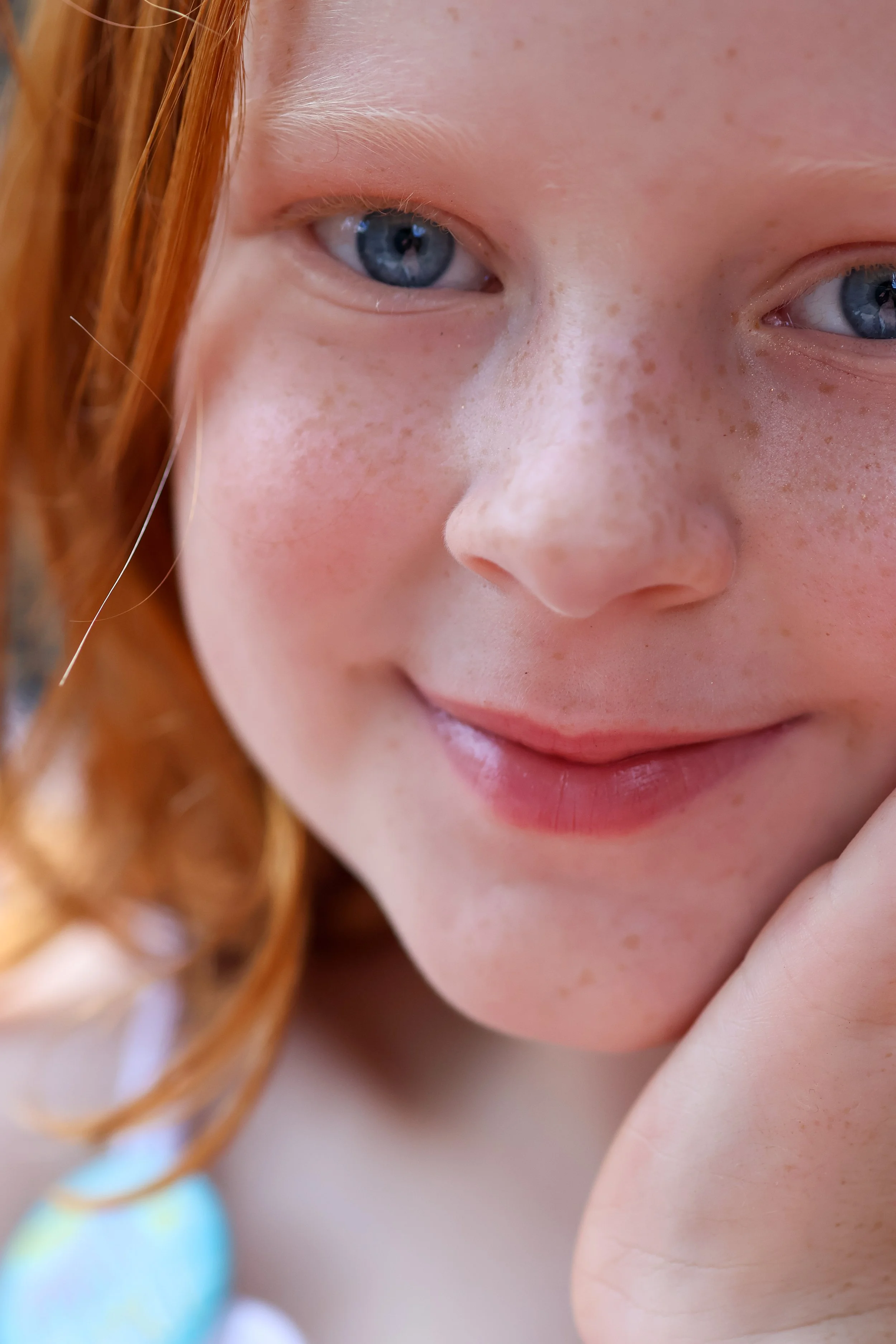 Close-up of a young person's face showing blue eyes, light freckles, and red hair. Their hand is near their chin, and the background is blurred.