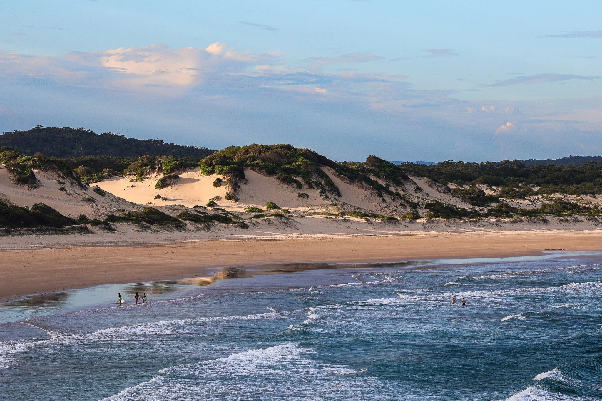 People walking along the beach shoreline with gentle waves, sandy dunes with sparse vegetation, and forested hills in the background under a partly cloudy sky.
