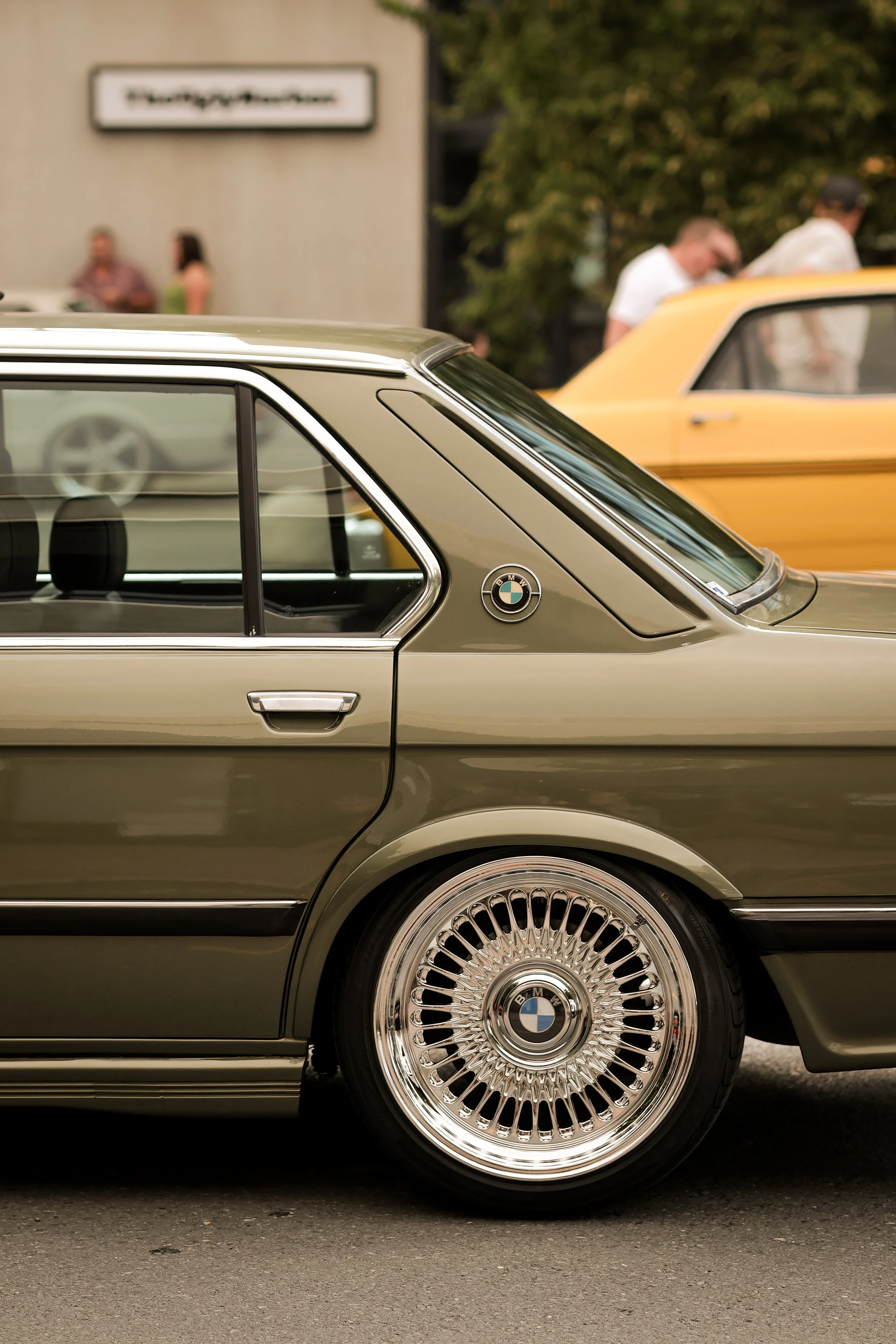 A close-up side view of a vintage BMW car with a chrome wire wheel, parked on a street. In the background, there are people and a building with a sign that is blurred.