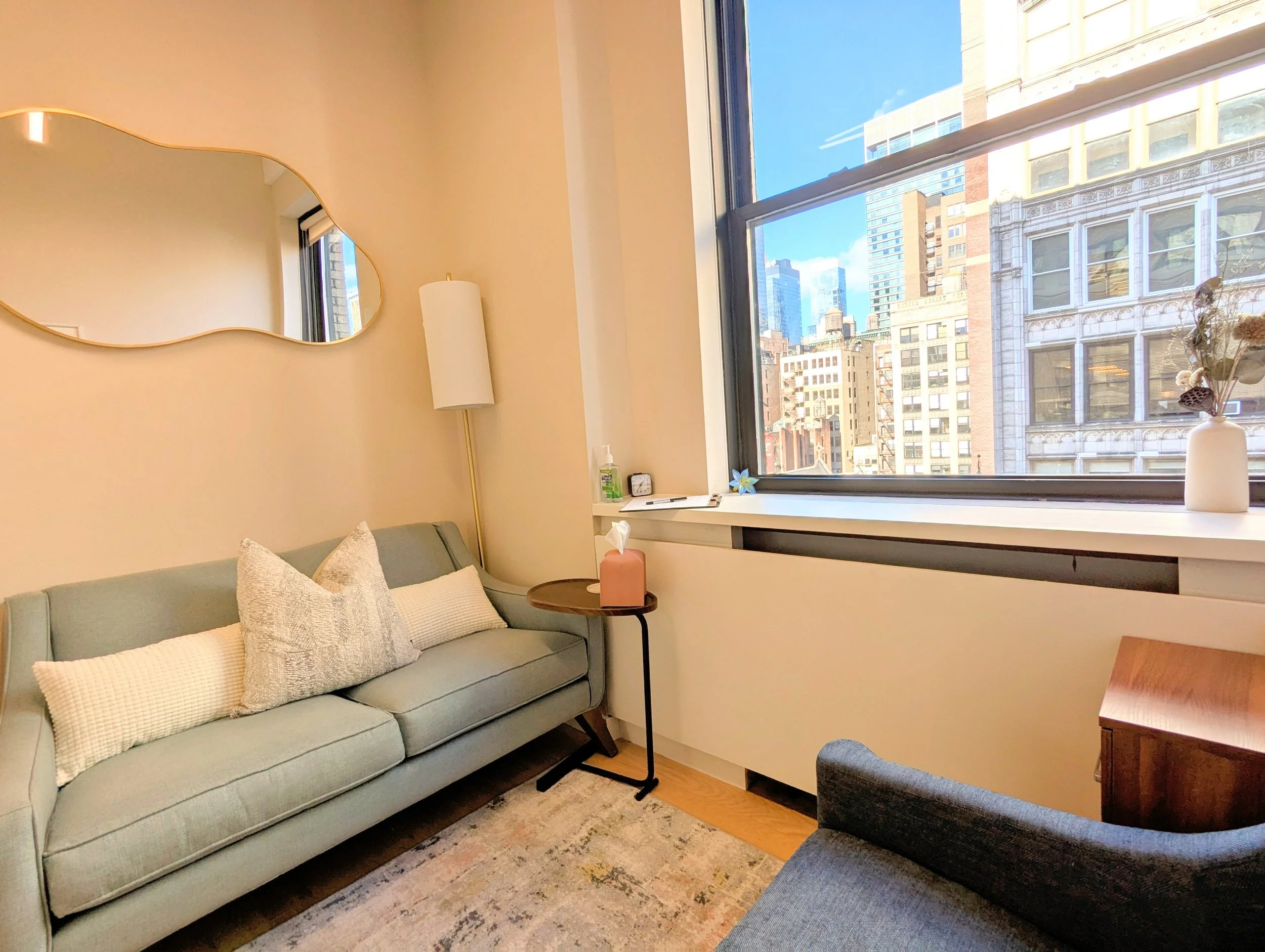 A cozy living room with two light-colored sofas, decorated with pillows. There is a large window showing city buildings and blue sky outside. A small round table next to the sofa holds a pink tissue box. A white vase with dried flowers sits on the windowsill, and a standing lamp is also visible.