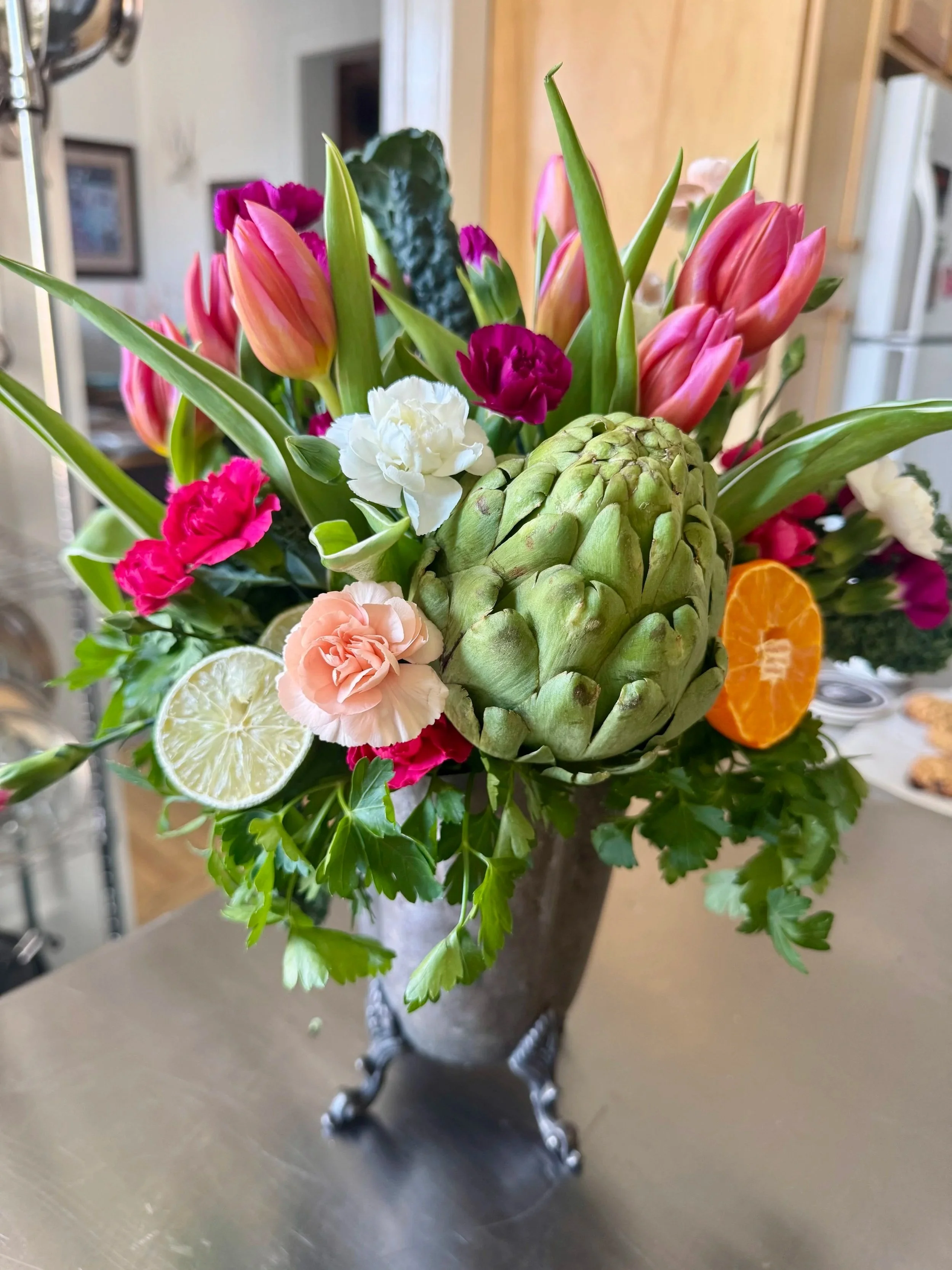 Bouquet of tulips, carnations, kale, an artichoke, and citrus slices in a silver footed vase