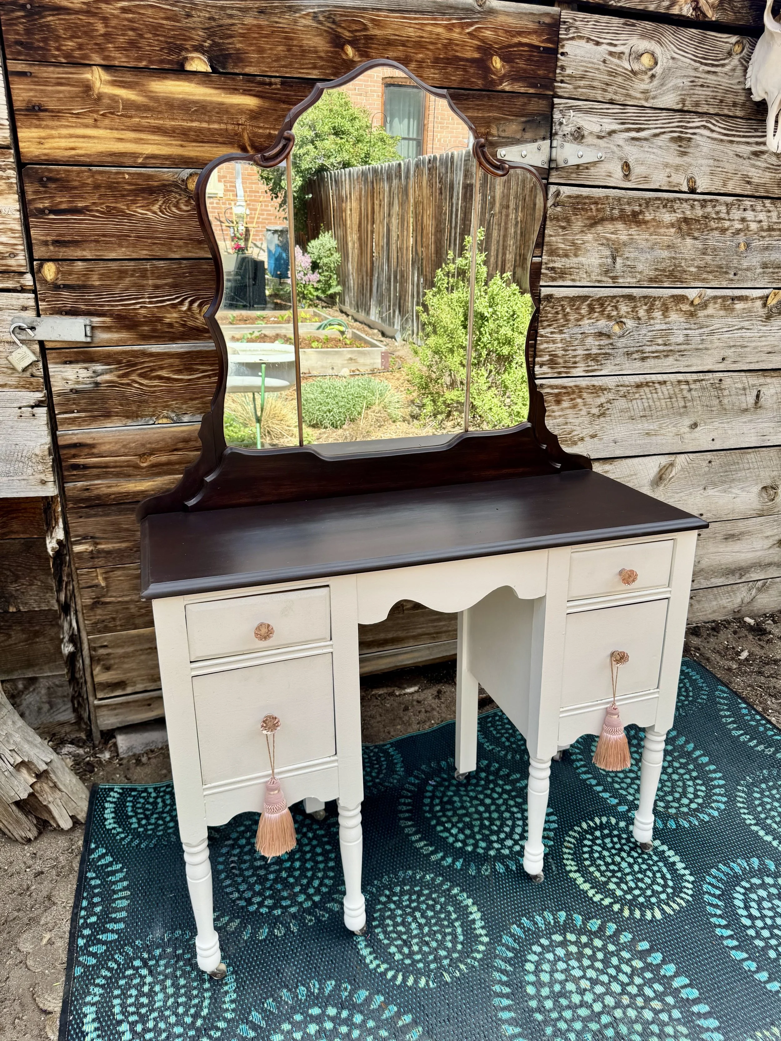 A vintage white vanity table with a dark wooden top, decorated with pink tassel drawer pulls, and a scalloped black mirror reflects a backyard garden with plants, a wooden fence, and a brick house in the background. The table is on a blue circular patterned rug.