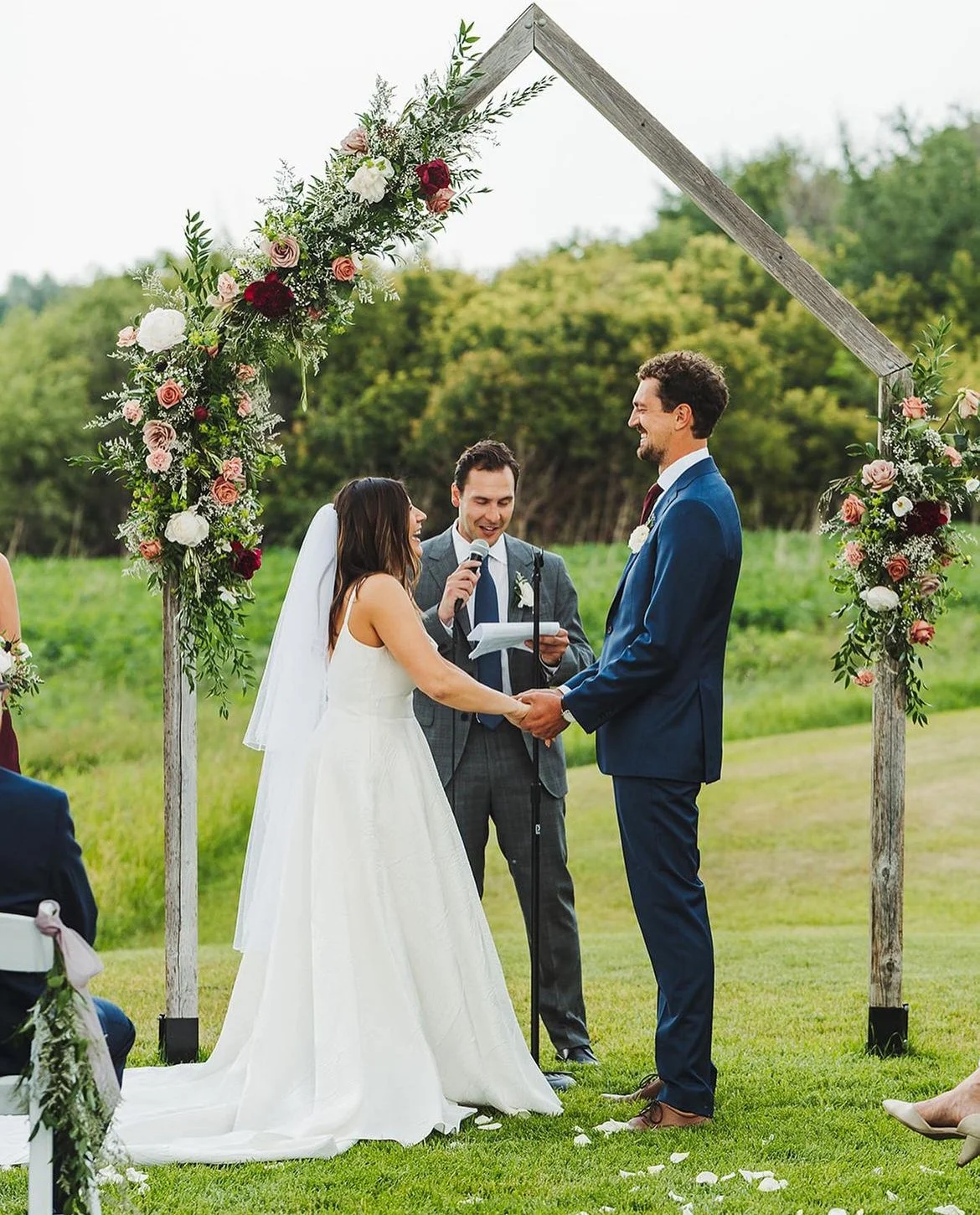 Bride and groom holding hands during an outdoor wedding ceremony with floral arch and officiant. Manitoba Marriage Commissioner.
