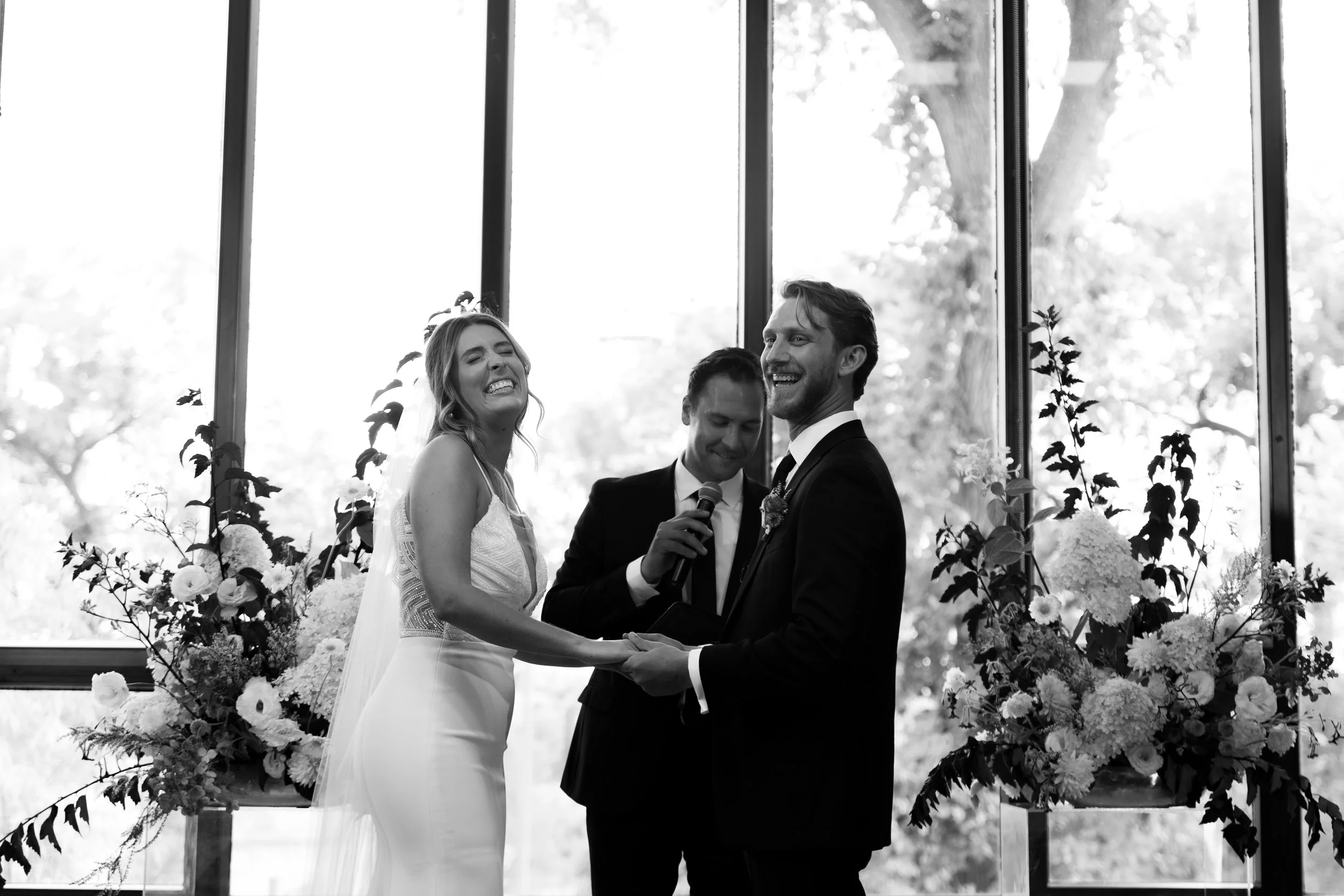 Manitoba Marriage Commissioner. Bride and groom holding hands during a wedding ceremony, smiling, with a man officiating in front of floral arrangements and large windows.