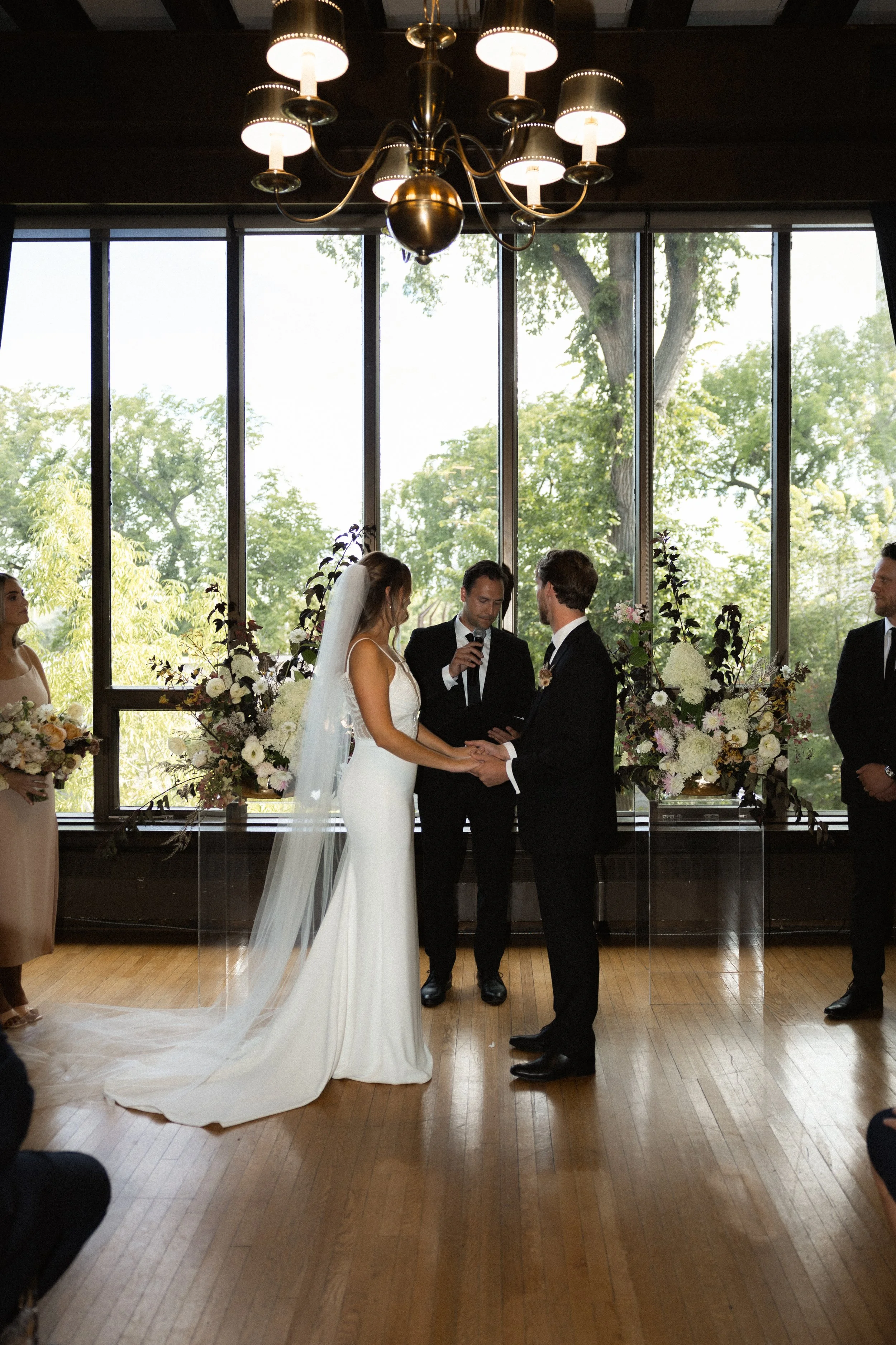 Manitoba Marriage Commissioner. Bride and groom exchanging vows during an indoor wedding ceremony with a officiant and attendants, surrounded by floral arrangements and large windows.