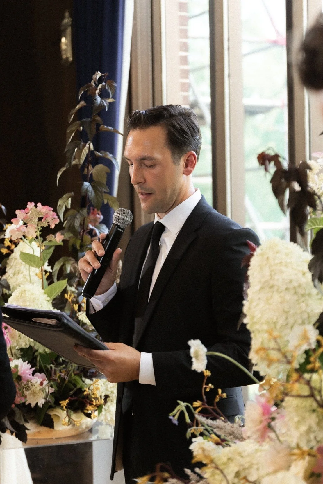 Man in a suit holding a microphone and reading from a folder, surrounded by floral decorations indoors.