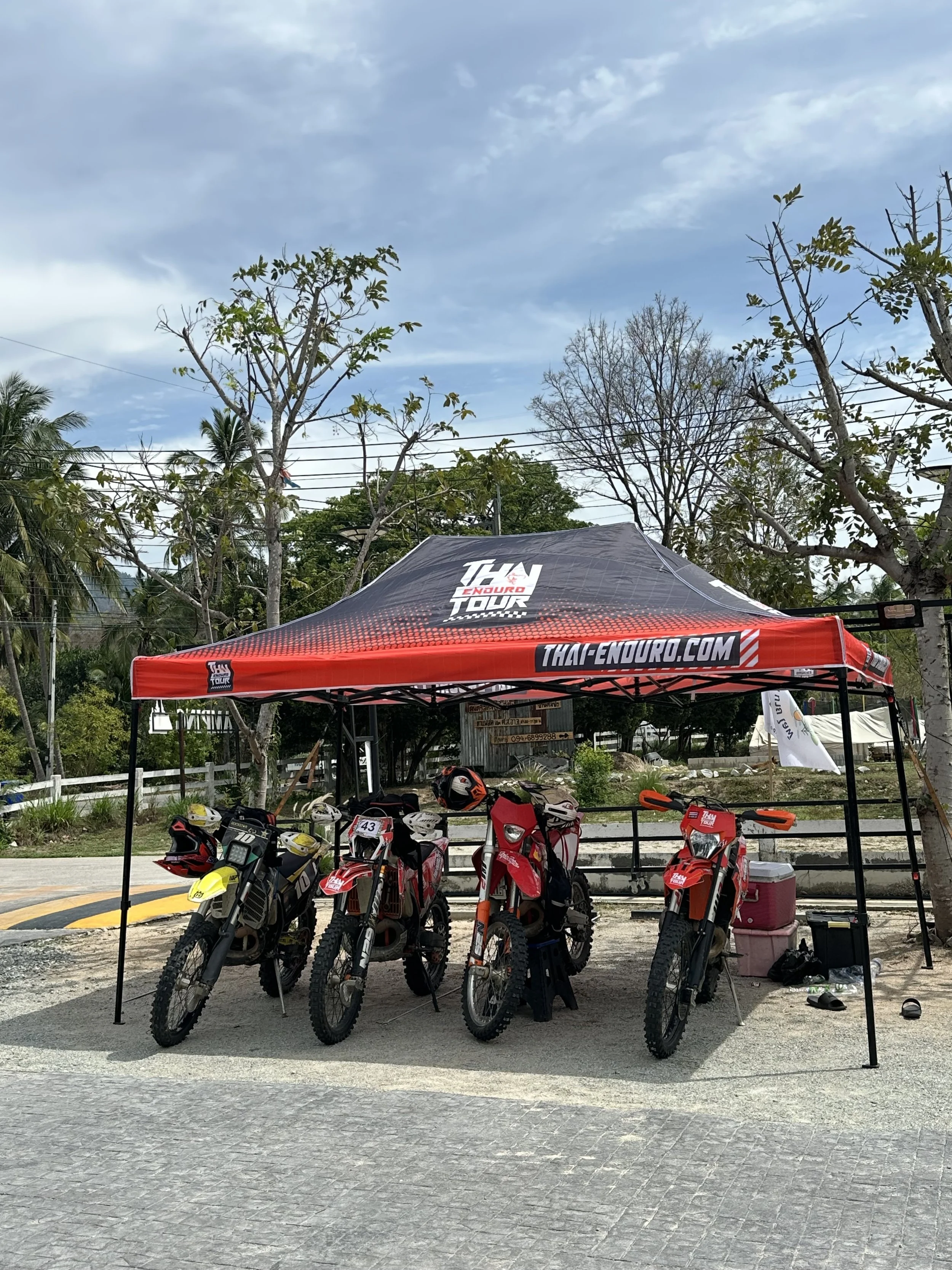 Four dirt bikes parked under a red and black canopy with 'Thai Enduro' branding, in an outdoor setting with trees and cloudy sky.