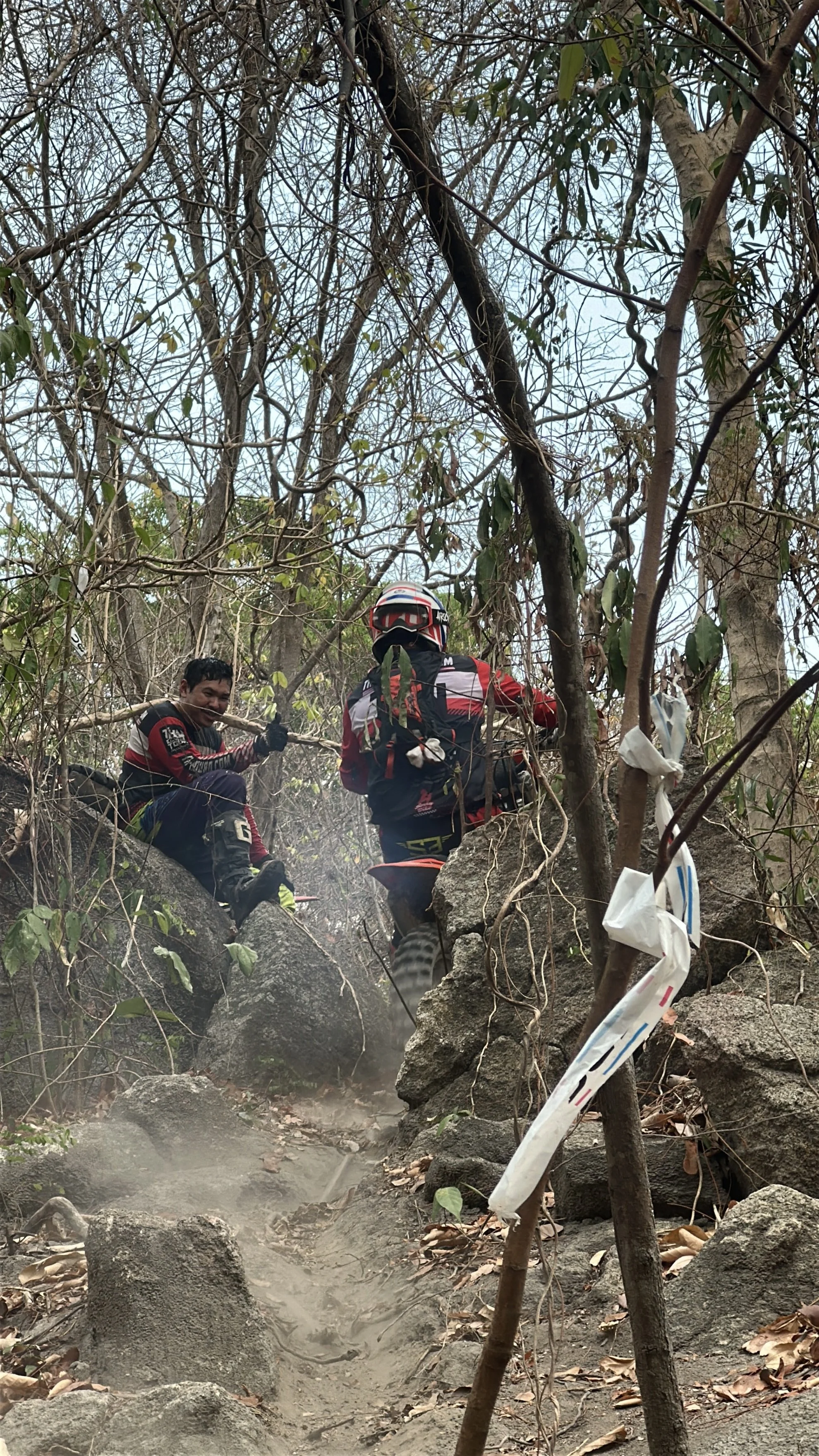 Two mountain bikers wearing helmets and backpacks navigating a rocky and dusty trail through a forested area with leafless and green trees.