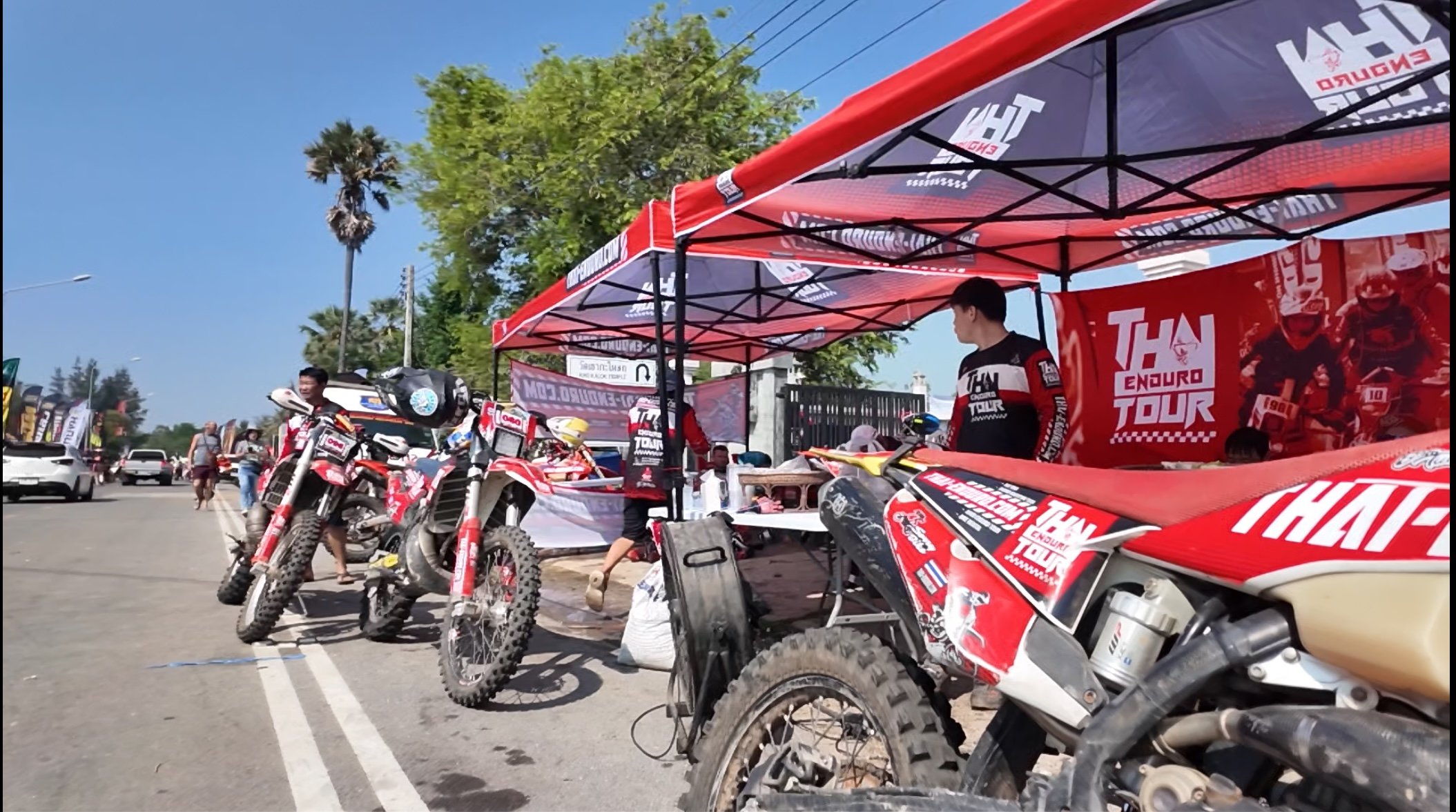Red and black dirt bikes parked under a canopy at an outdoor event, with people walking nearby and a large banner promoting THAI ENDURO TOUR in the background, on a sunny day with blue skies and green trees.