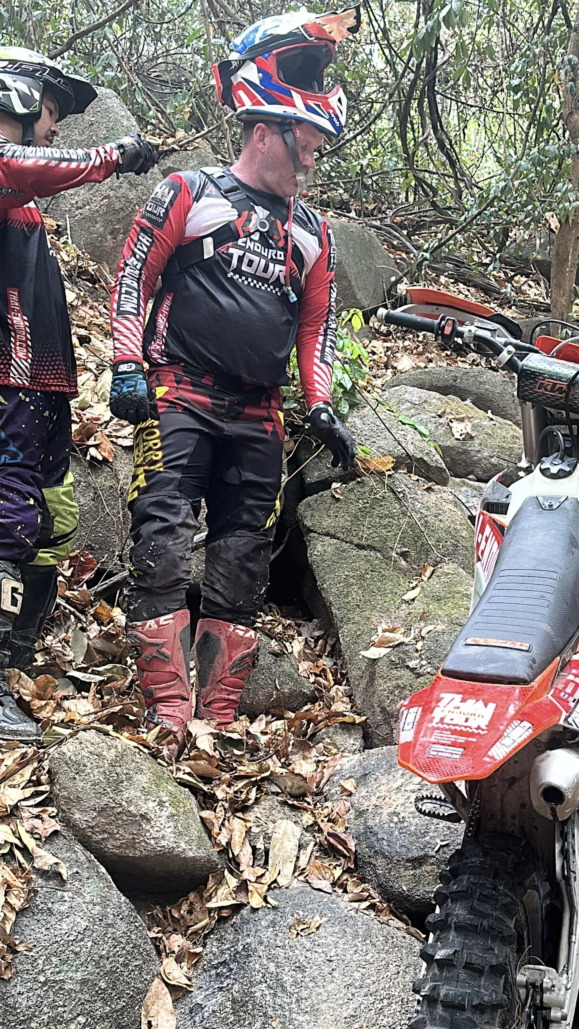 Two mountain bikers in riding gear navigate a rocky, wooded trail with their dirt bikes. One rider looks down, and they are surrounded by fallen leaves and large rocks.