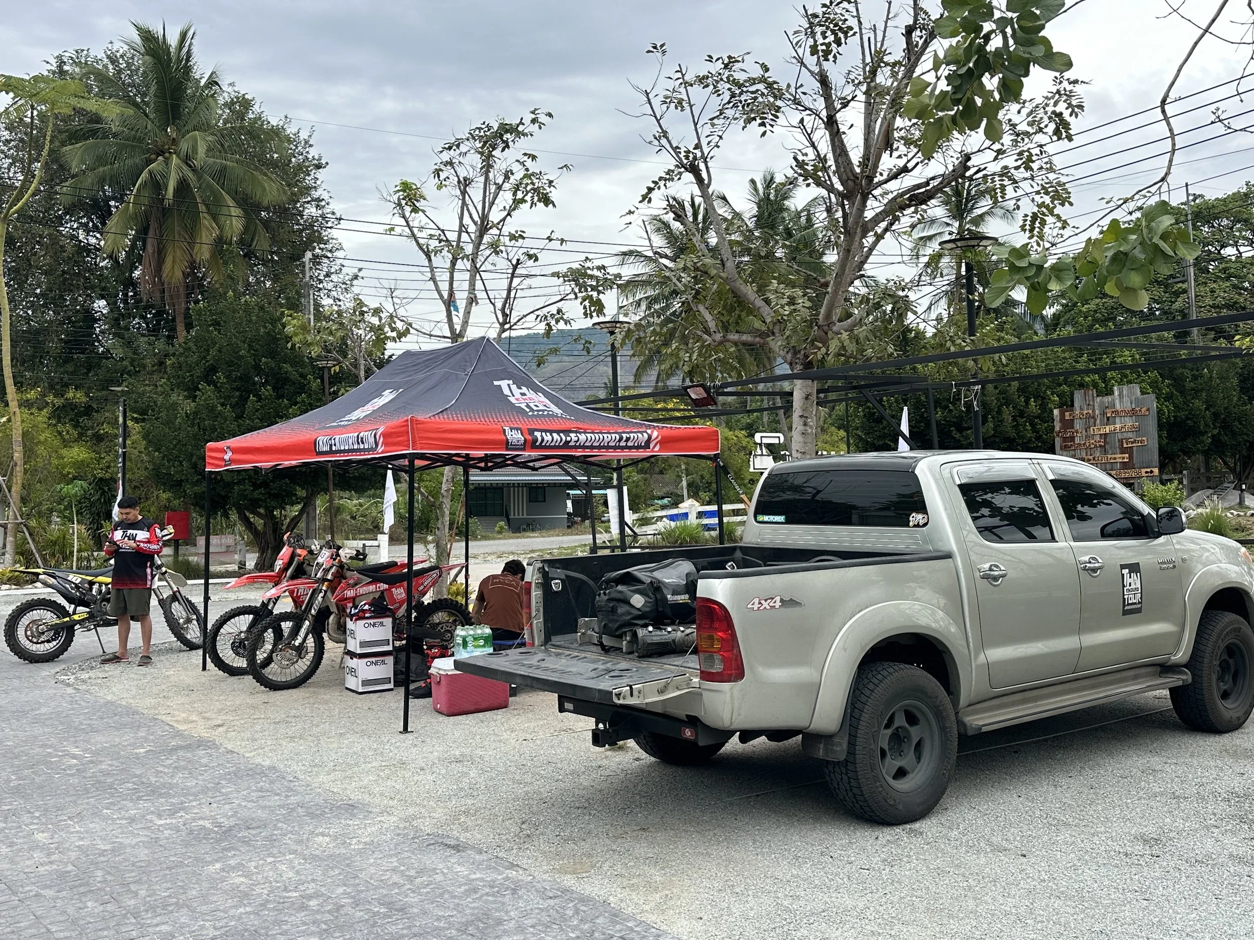 An outdoor setup with a red and black canopy tent, two dirt bikes, a person standing near a dirt bike, a person sitting under the tent, and a silver pickup truck parked nearby. The scene is surrounded by trees and electrical wires.