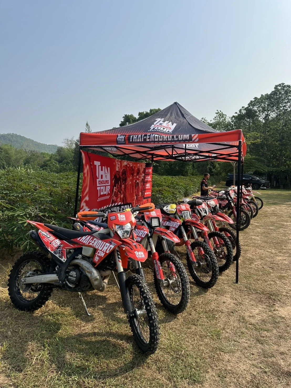 Thai Enduro Tour red dirt bike lineup parked beside tour tent with mountain and jungle scenery in Thailand