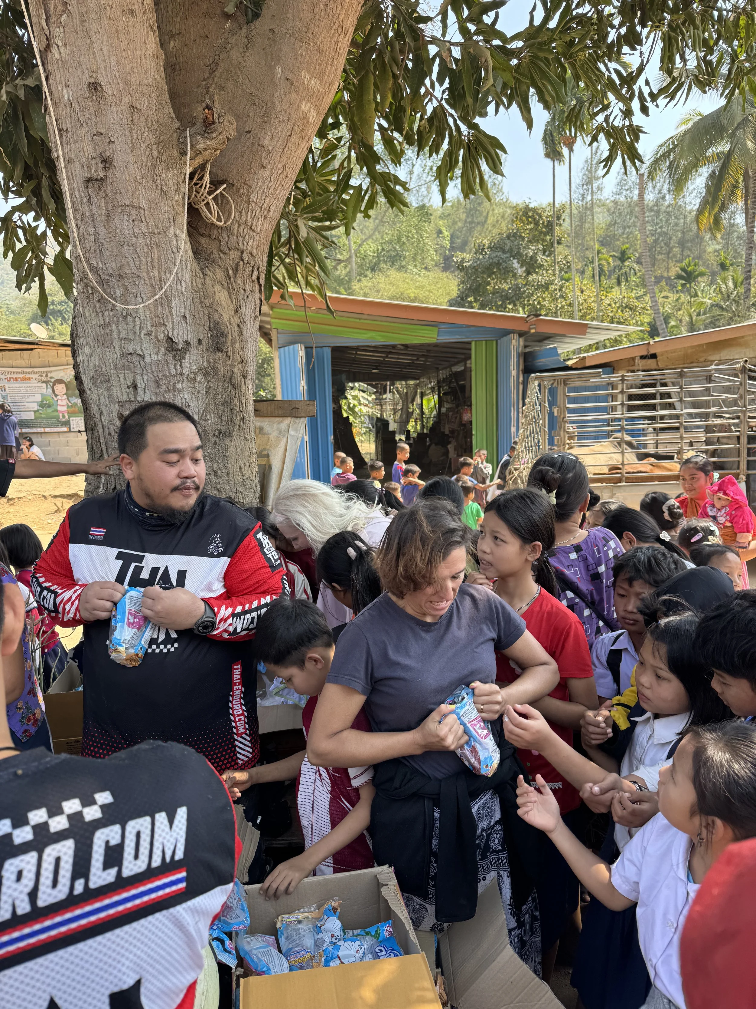 Enduro Tour Thailand riders distributing snacks and supplies to children under large tree in Thai countryside