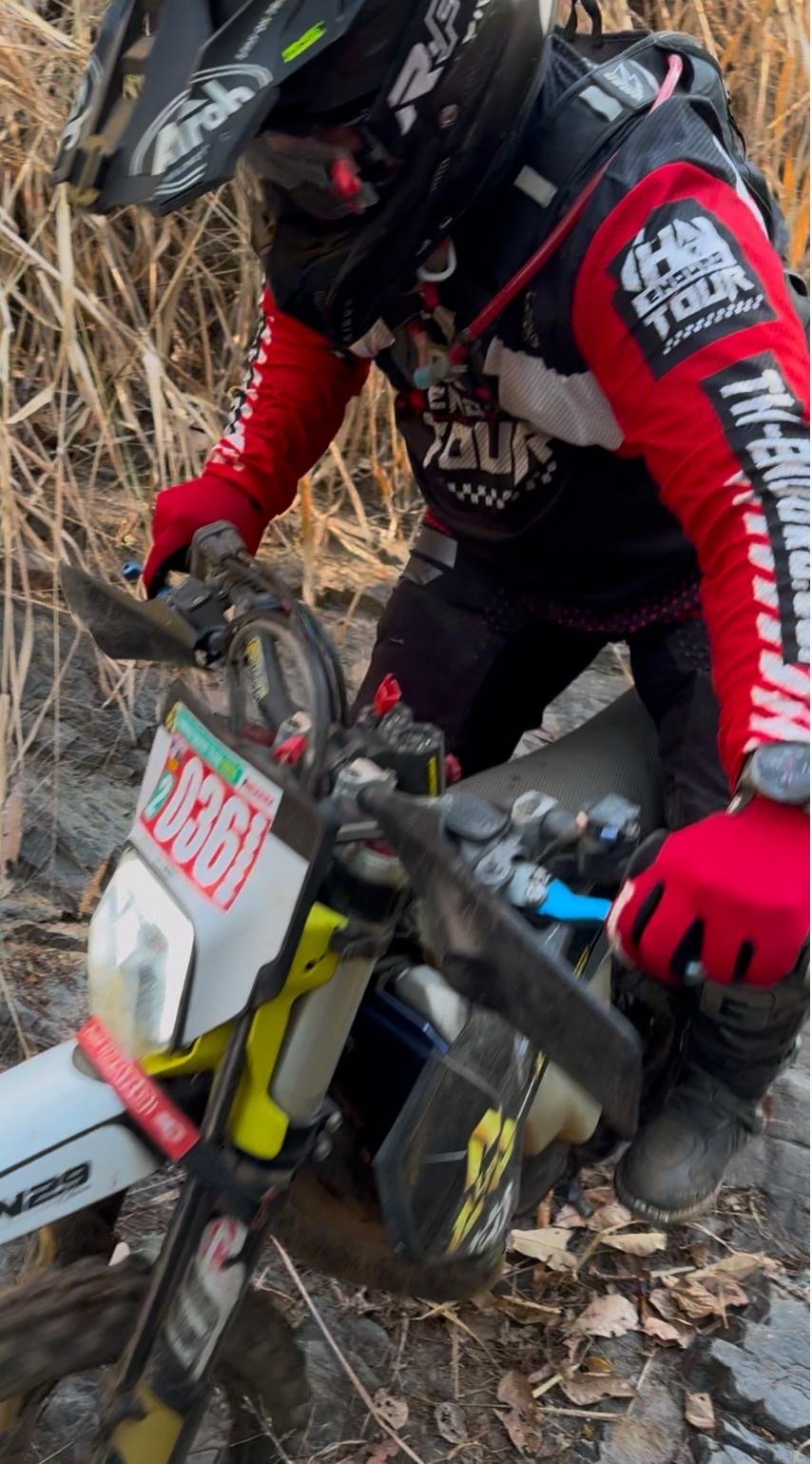 A person in riding gear, including a helmet and a red and black jacket with rider logo, riding a dirt bike through a forested area with dry branches and leaves on the ground.