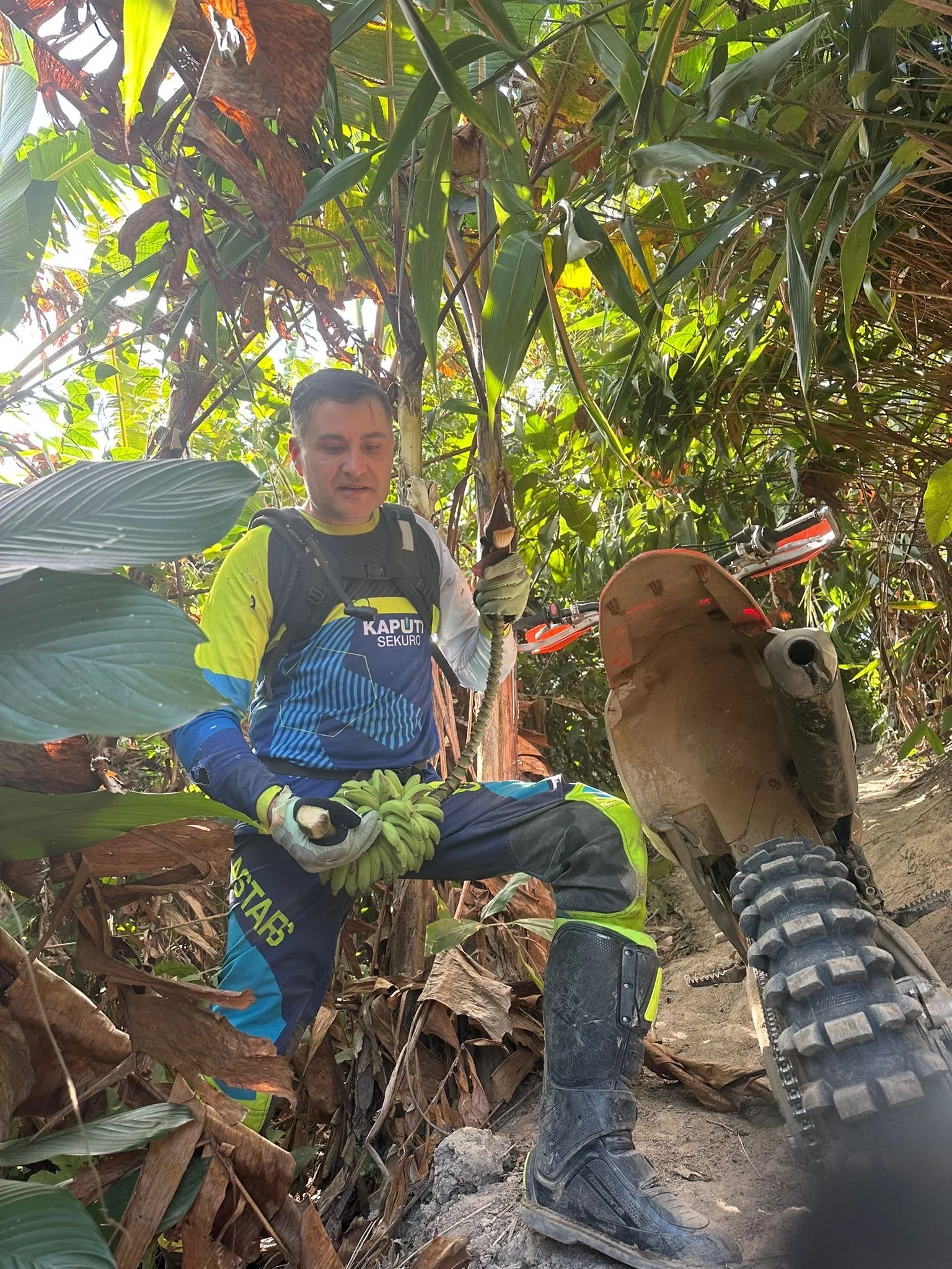 A man dressed in motocross gear, including a colorful jersey and pants, is sitting beside a dirt bike on a trail in a dense, leafy jungle. He is holding a snake and appears to be inspecting or managing it, surrounded by green vegetation.