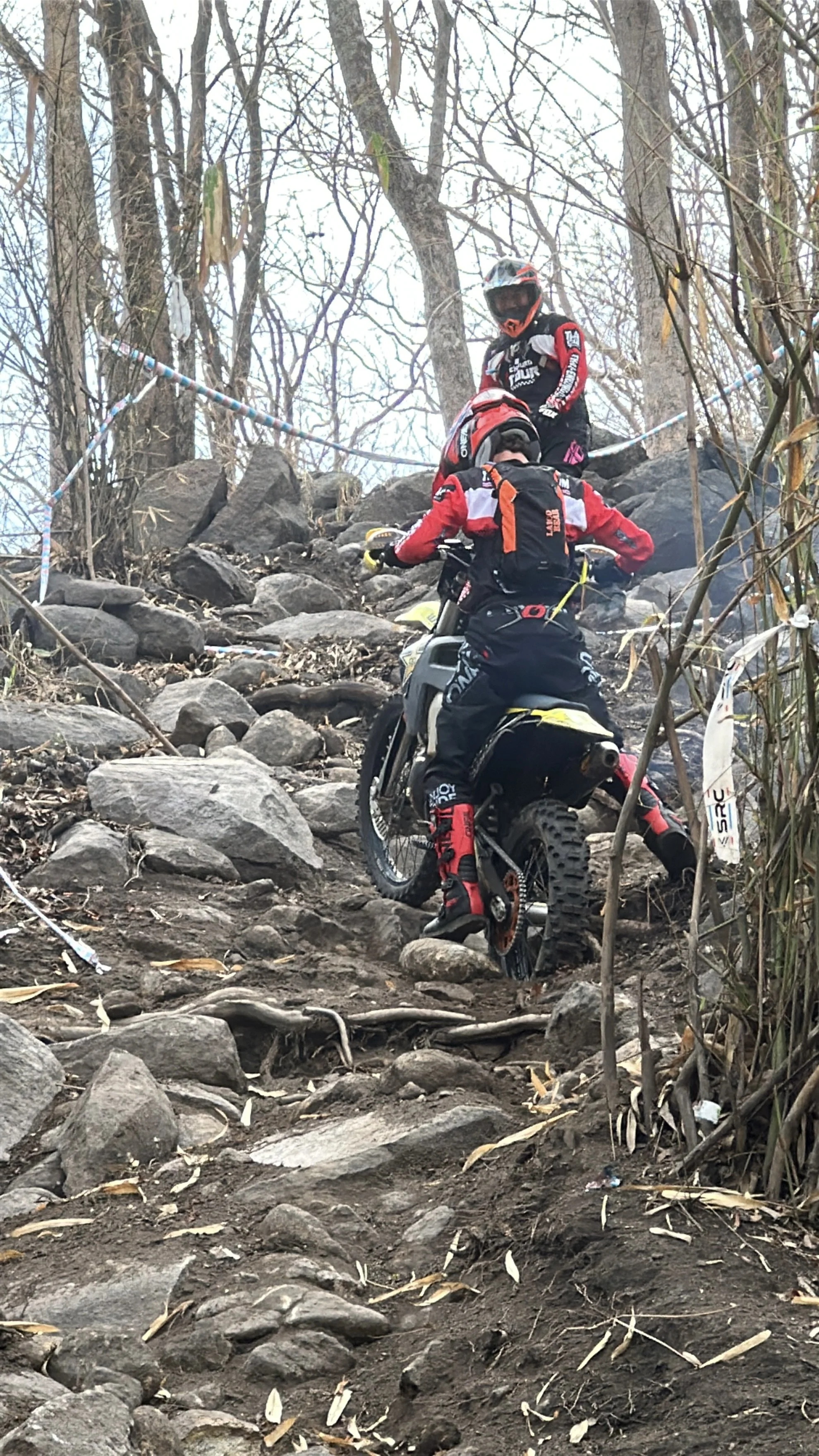 Two children wearing mountain biking gear, including helmets and protective clothing, climb a rocky trail in a wooded area, with trail ropes on either side.