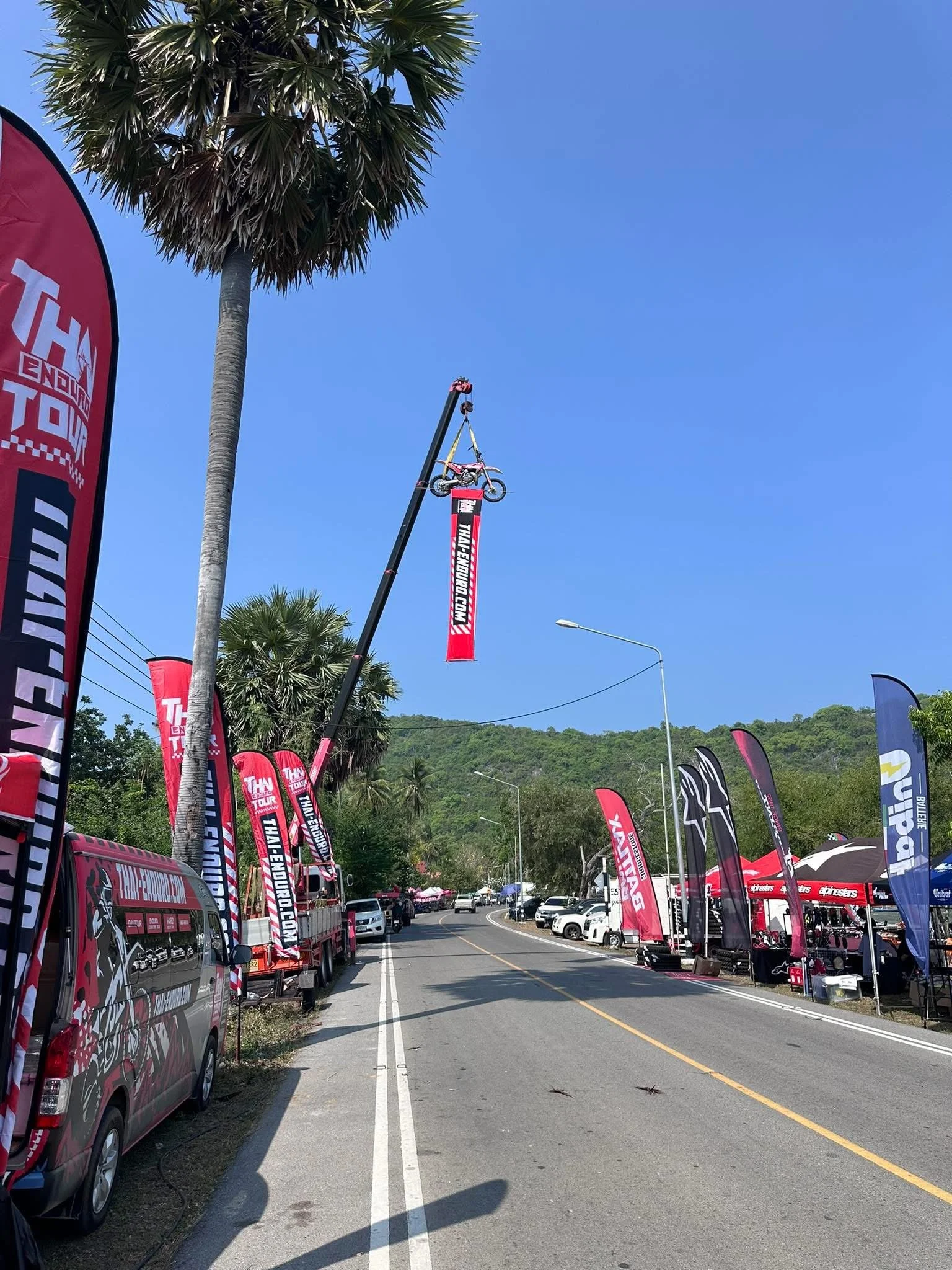 A mountain biking event setup on a sunny day, featuring flags and banners with the event branding, parked cars, tents, and a mountain in the background.
