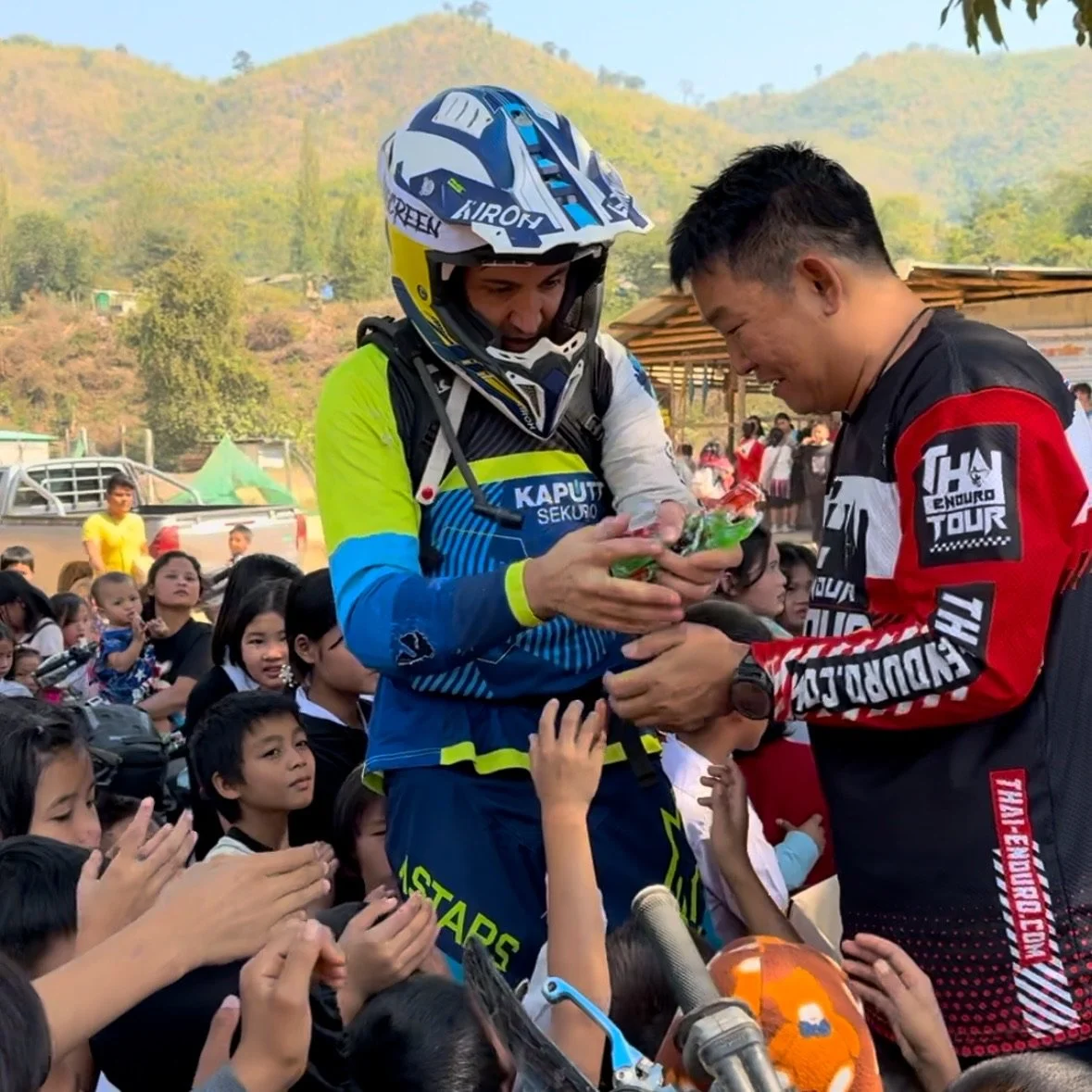 Enduro motorcycle rider receiving a gift during a Thailand enduro tour event, with spectators, children, hills, and forest landscape in the background