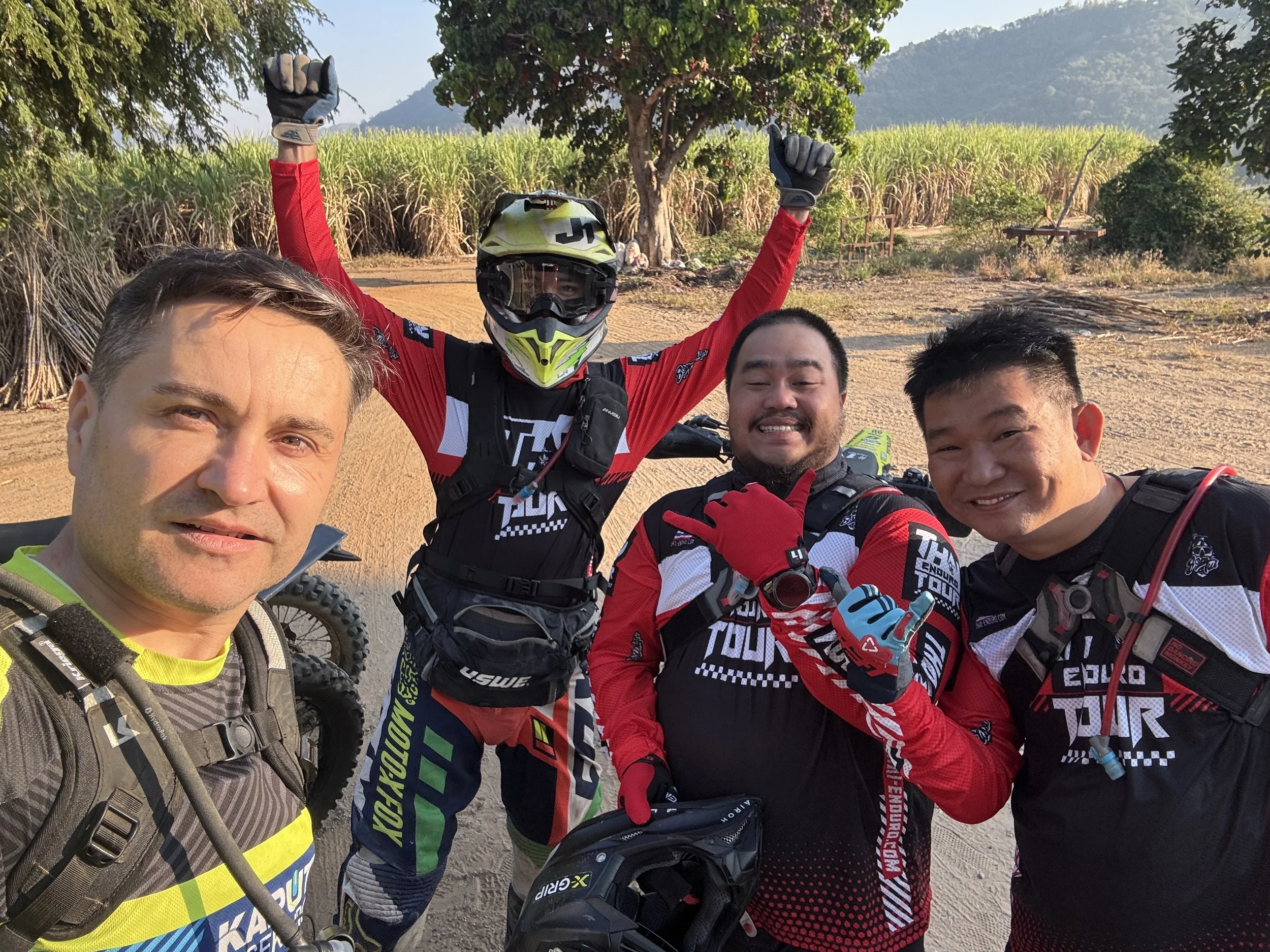 Group of enduro riders taking a selfie on a dirt trail in Thailand, surrounded by lush green jungle, mountain scenery, and off-road adventure landscape