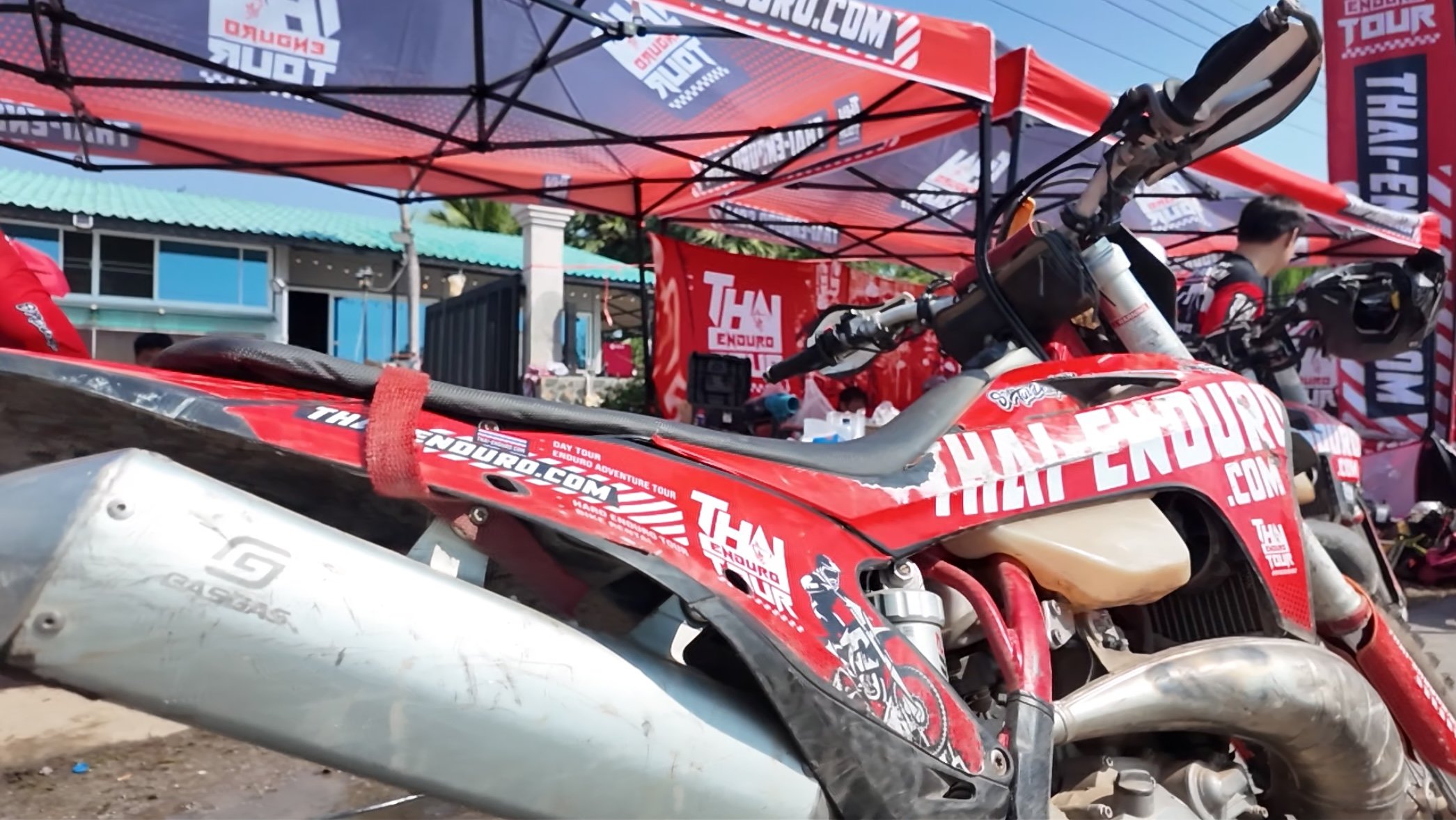 A red and black dirt bike with decals reading 'THAN ENDURO' and 'ENDURO.COM' under a red canopy, with a person standing in the background.