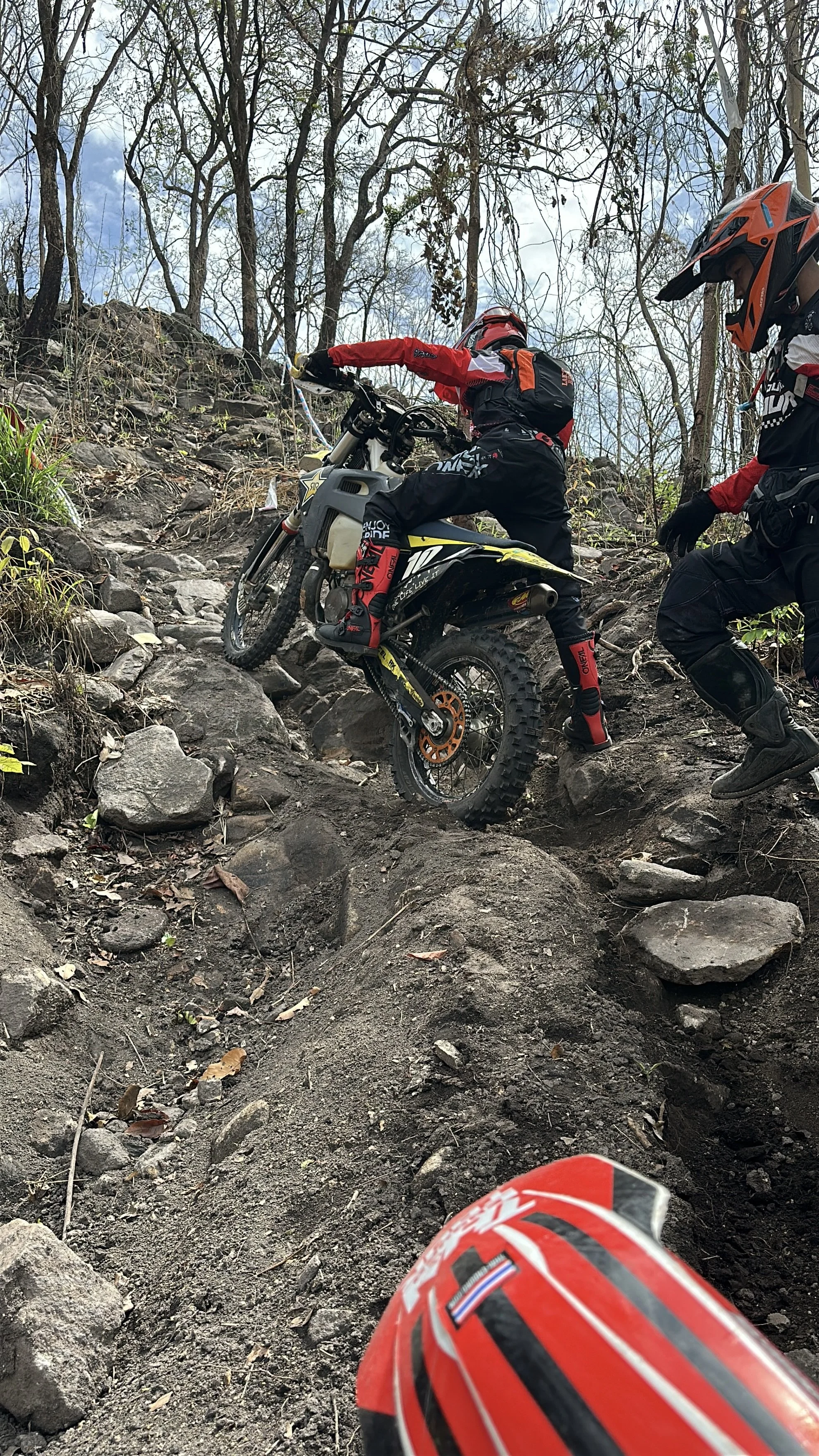 Two dirt bike riders climbing a rocky, steep trail through a forest with leafless trees and a partly cloudy sky.