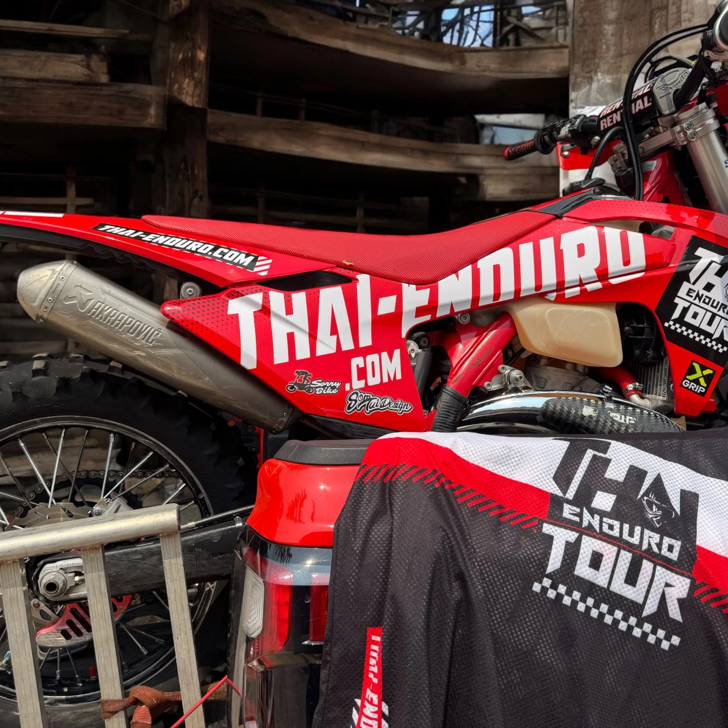 Close-up of a red and black motocross bike with multiple stickers, including 'THAI-ENDURO.COM' and 'TRA
dENDURO.COM'. The bike is parked next to a red and black vehicle or equipment with similar branding, and a part of a tire and a metallic structure