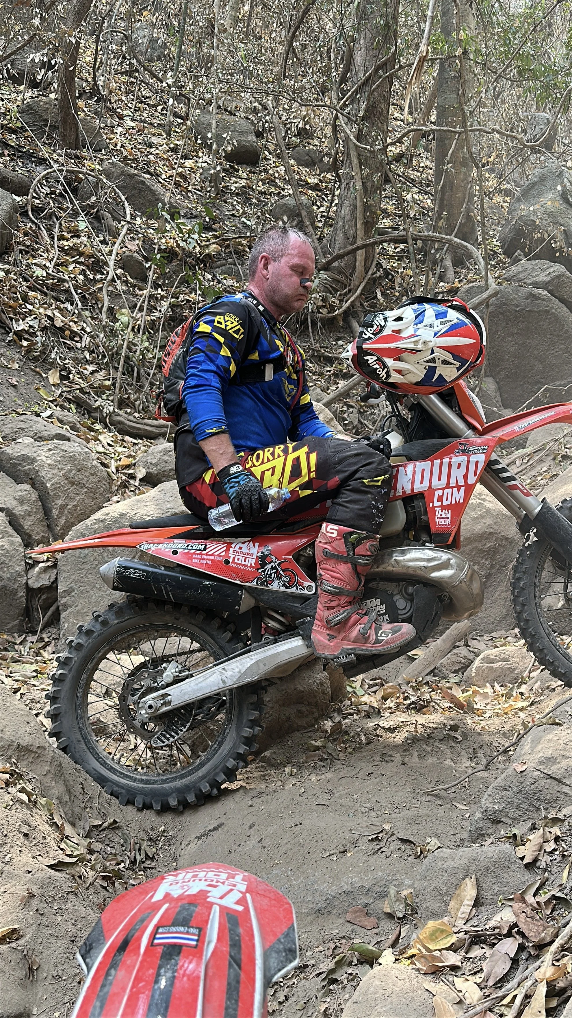 A motocross rider wearing a blue and black outfit sitting on a red dirt bike amidst a wooded trail, holding a water bottle, with a helmet on the bike's handlebars.