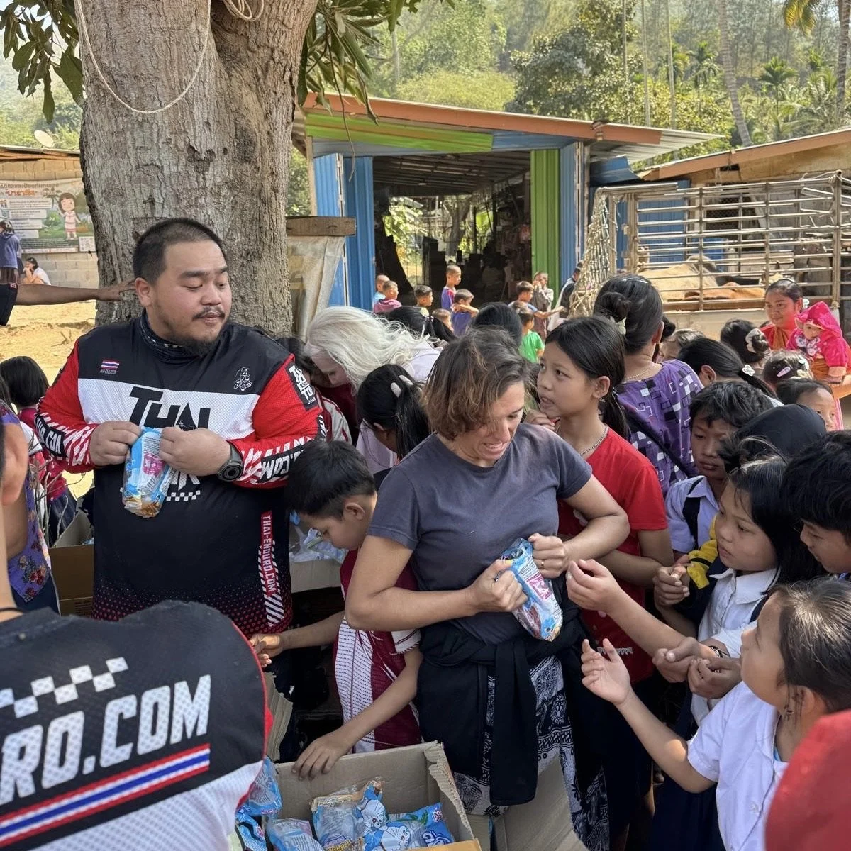 Enduro Tour Thailand riders giving snacks to local children during an enduro community activity in rural Thailand, with dirt bikes and nature in the background — adventure motorcycle charity moment
