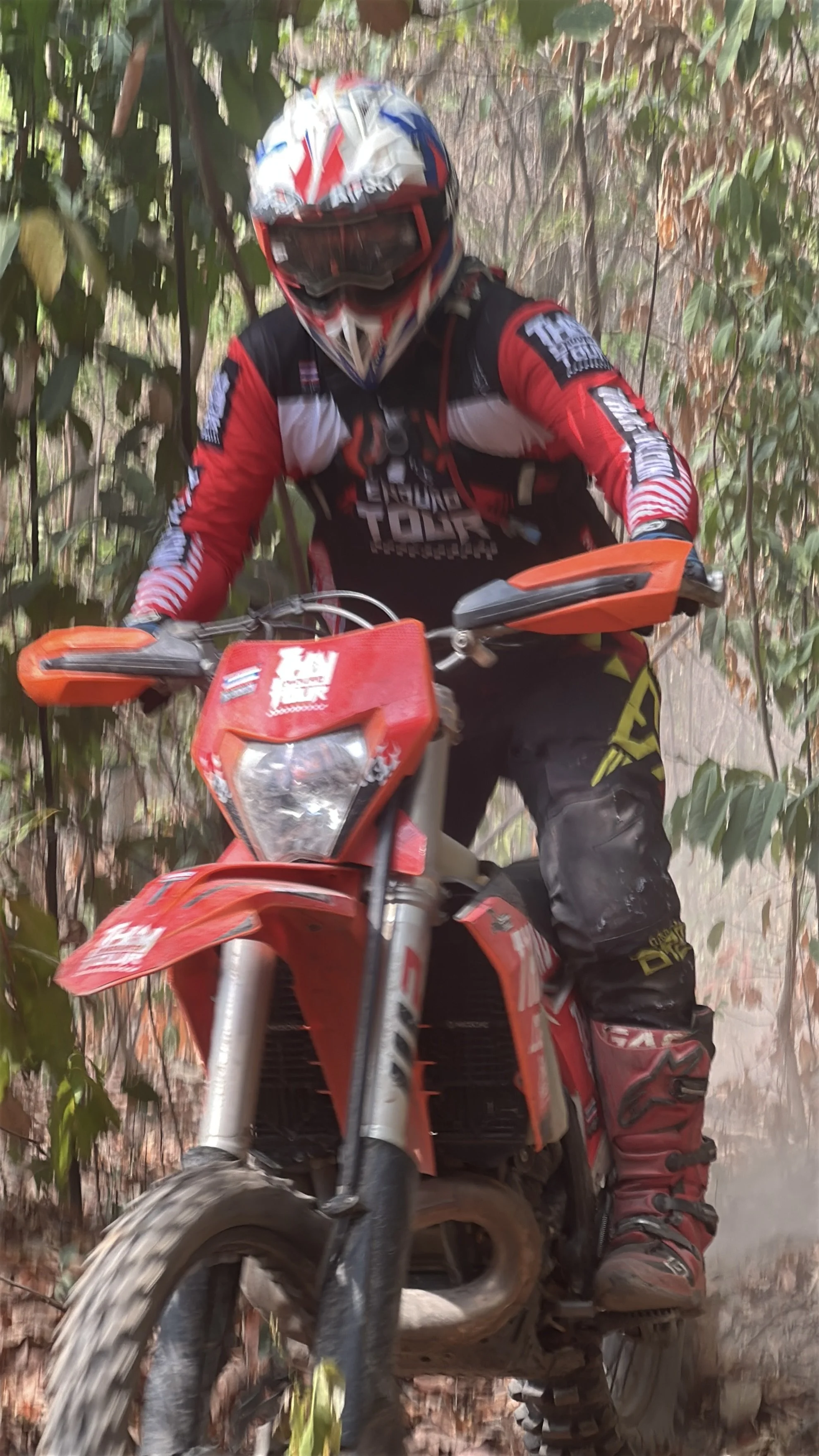 A motocross rider wearing a helmet, goggles, and riding gear navigates through a narrow dirt trail surrounded by foliage on a red dirt bike.