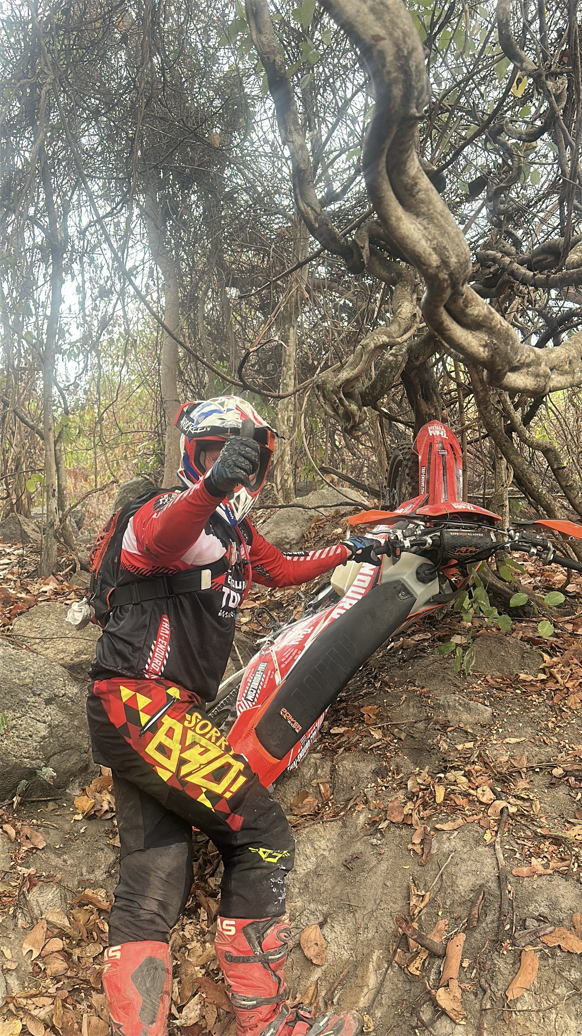 A dirt bike rider in full gear giving a thumbs-up, standing next to an orange and white off-road motorcycle on rocky terrain in a wooded area.