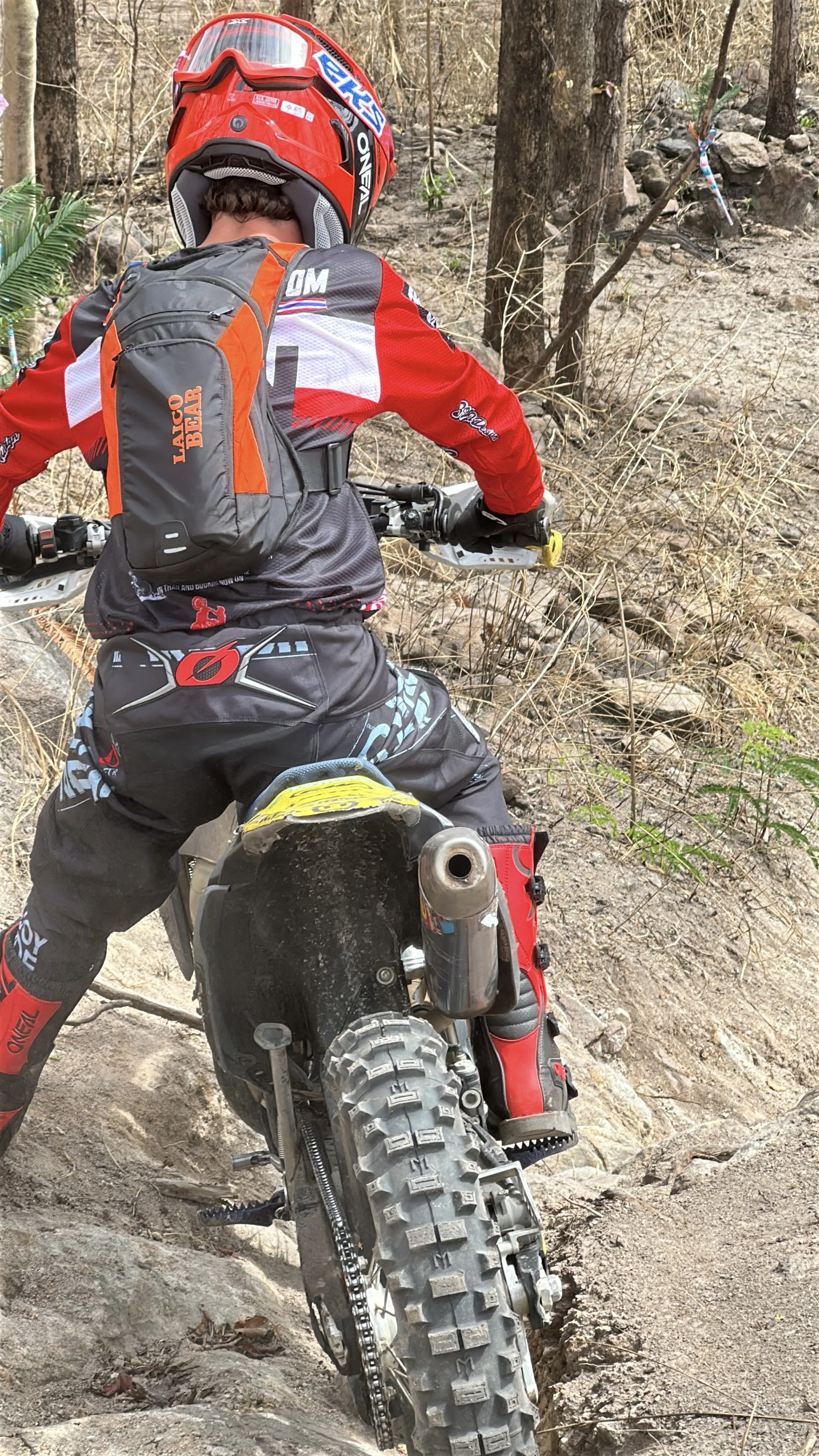 Motocross rider wearing a red helmet and protective gear navigating rocky terrain in a forest.