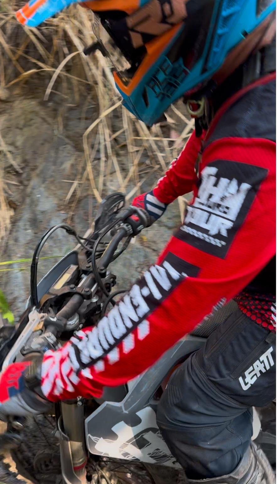 A person riding a dirt bike on a trail through a wooded area with tall dry grass and trees.