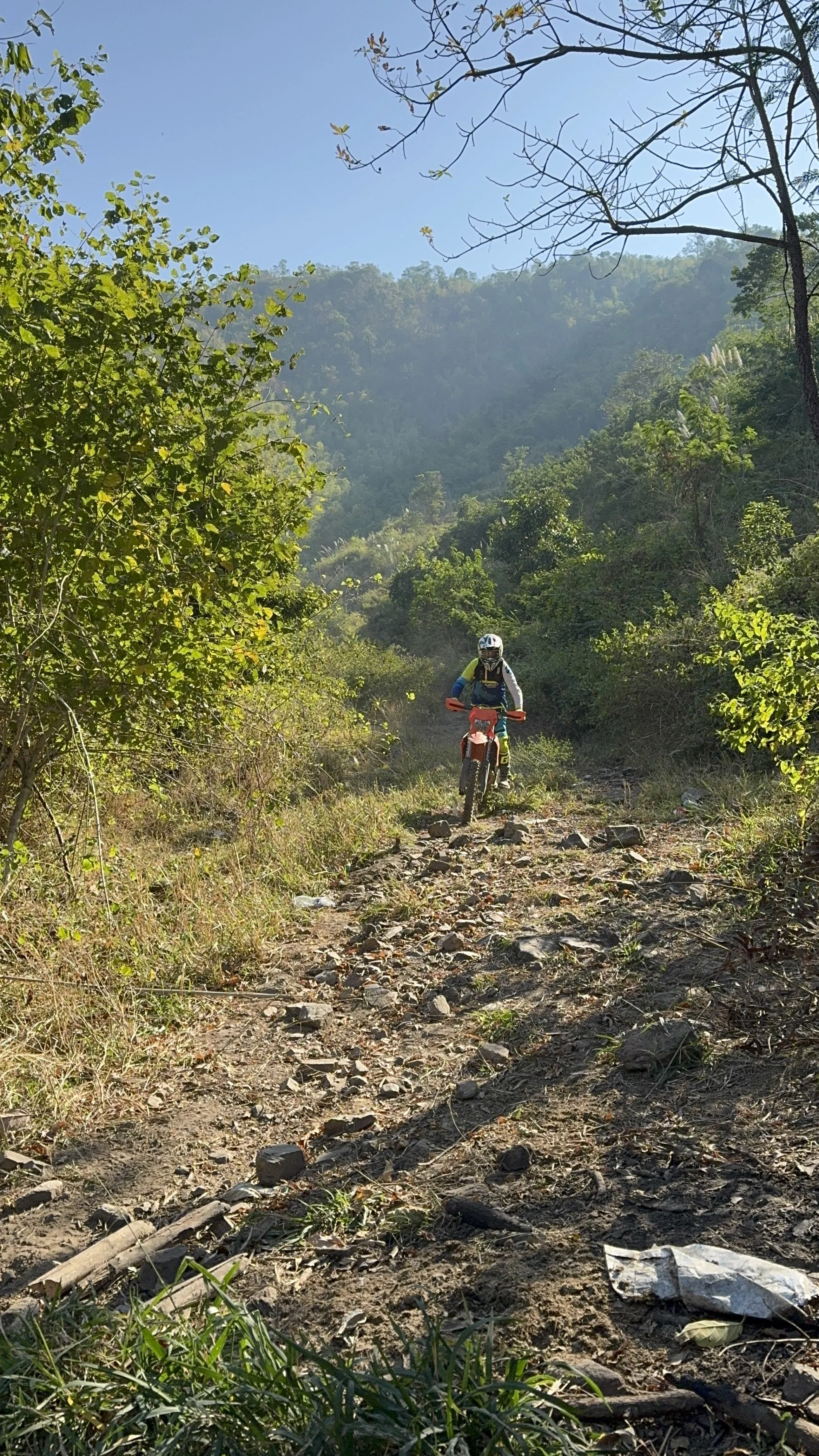 Enduro rider navigating a rocky uphill dirt trail in a jungle mountain area of Northern Thailand under clear blue sky — off-road enduro tour experience