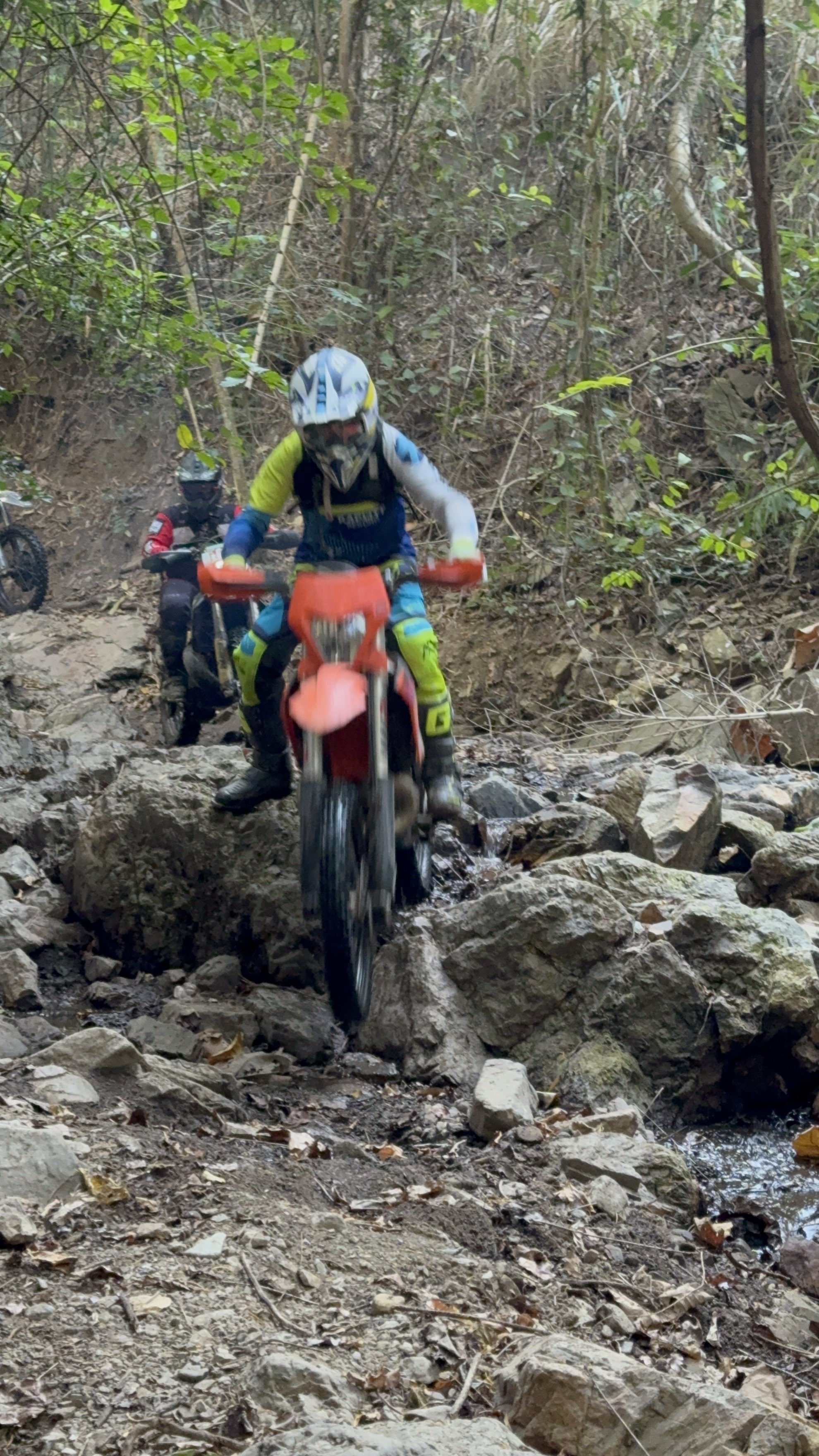 oung rider on a small dirt bike crossing rocks and shallow water on a forest trail — family-friendly enduro riding in Thailand