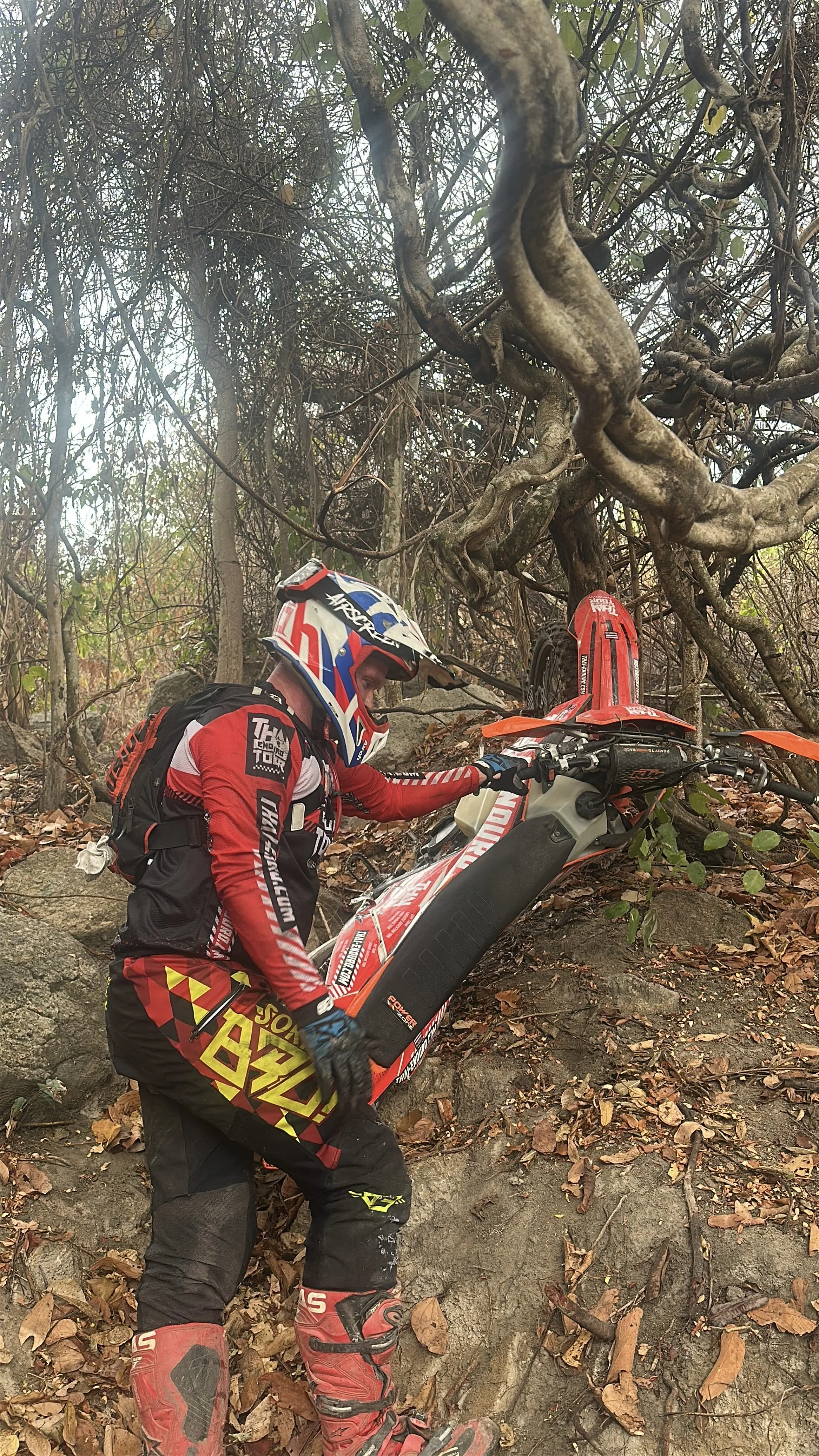 A person in motocross gear, including a red, white, and blue helmet and red and black protective suit, is climbing a rocky, leaf-covered hill while trying to lift a red and white dirt bike with orange accents.