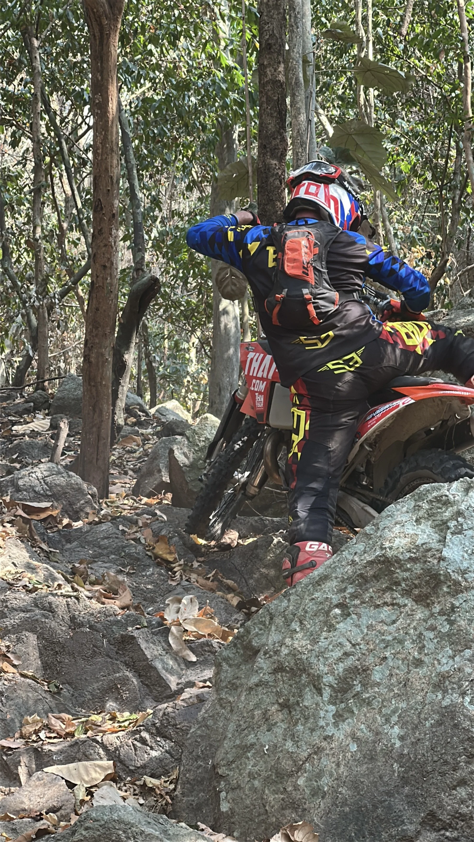 A person riding a dirt bike through a rocky forest trail, wearing a helmet, long-sleeve jersey, and riding gear.