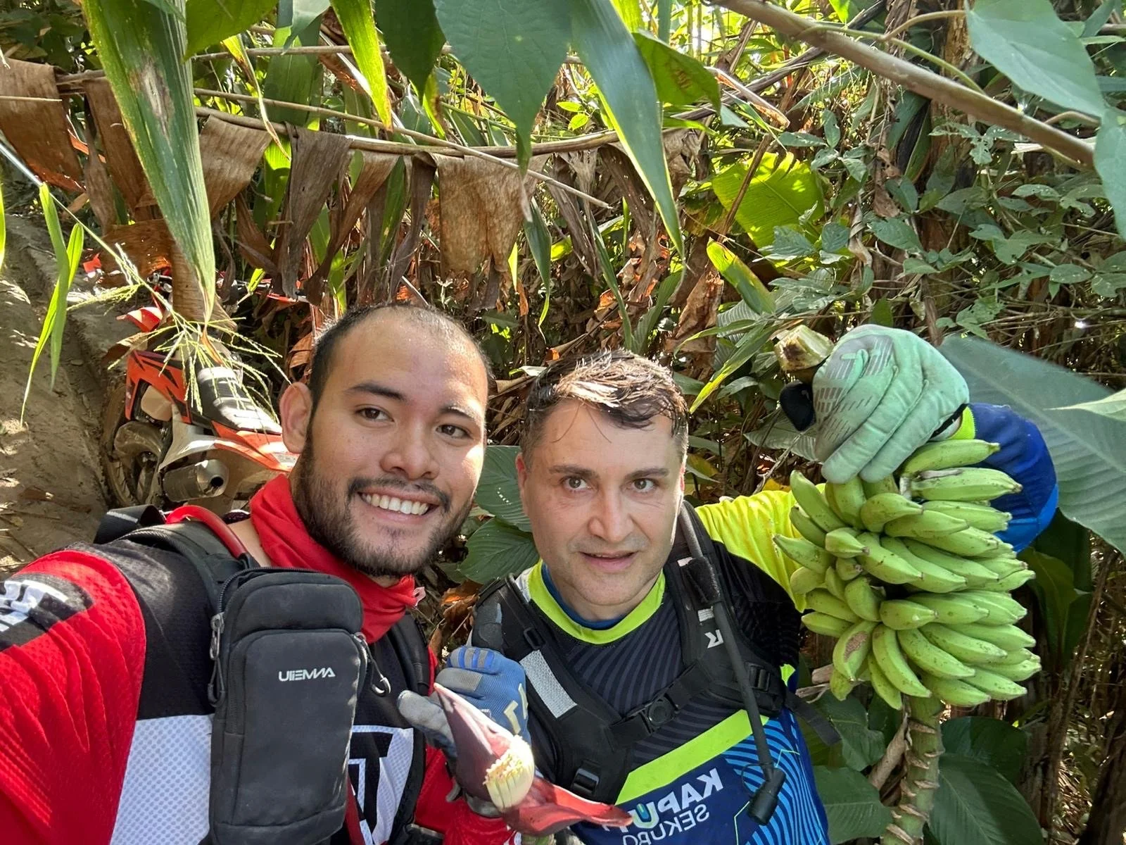 Two men in motorcycle gear and gloves standing in a jungle next to a banana plant, holding a bunch of bananas and smiling at the camera.