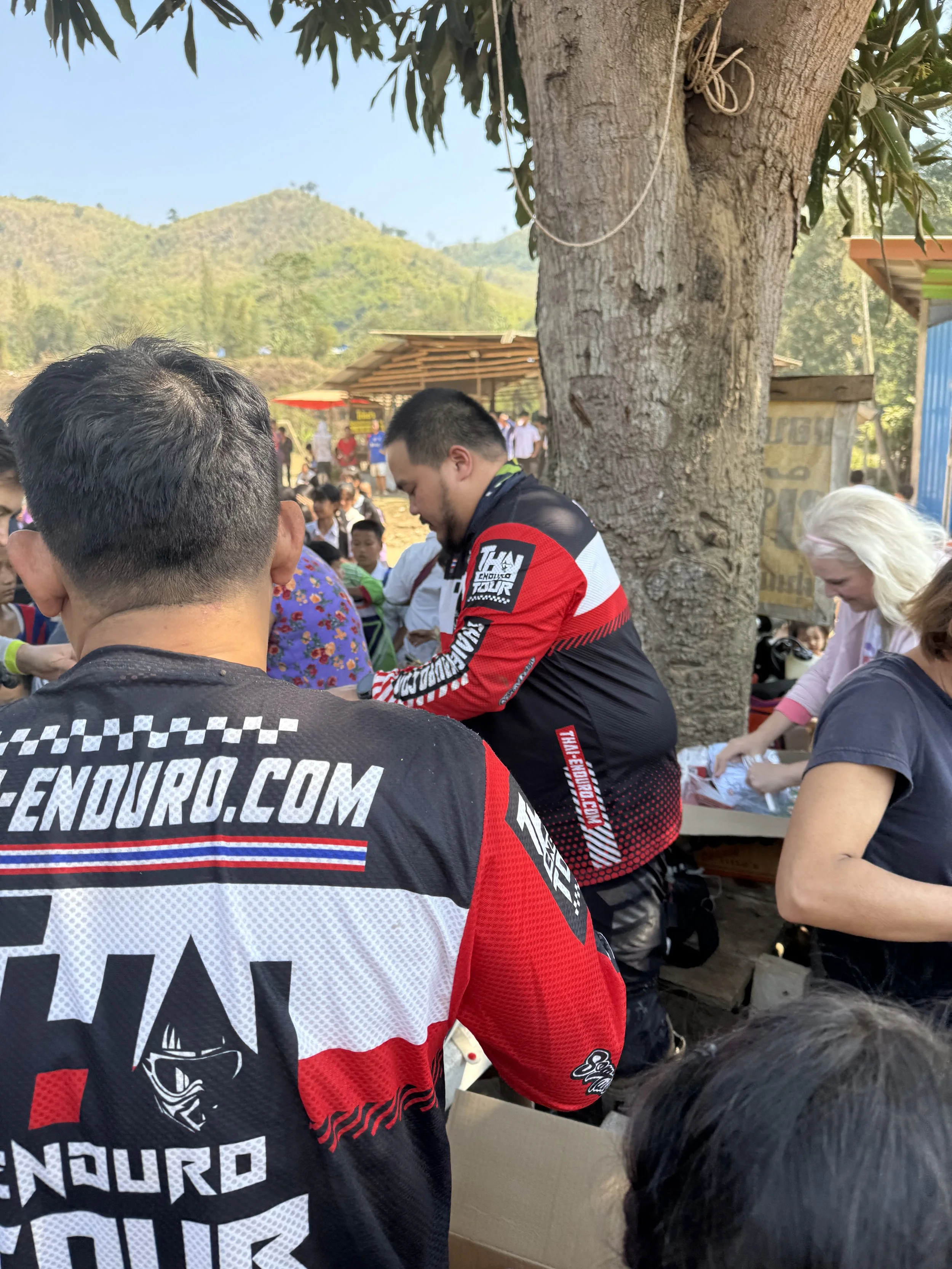 Enduro Tour Thailand community event with riders and locals gathering under large tree in rural mountain area