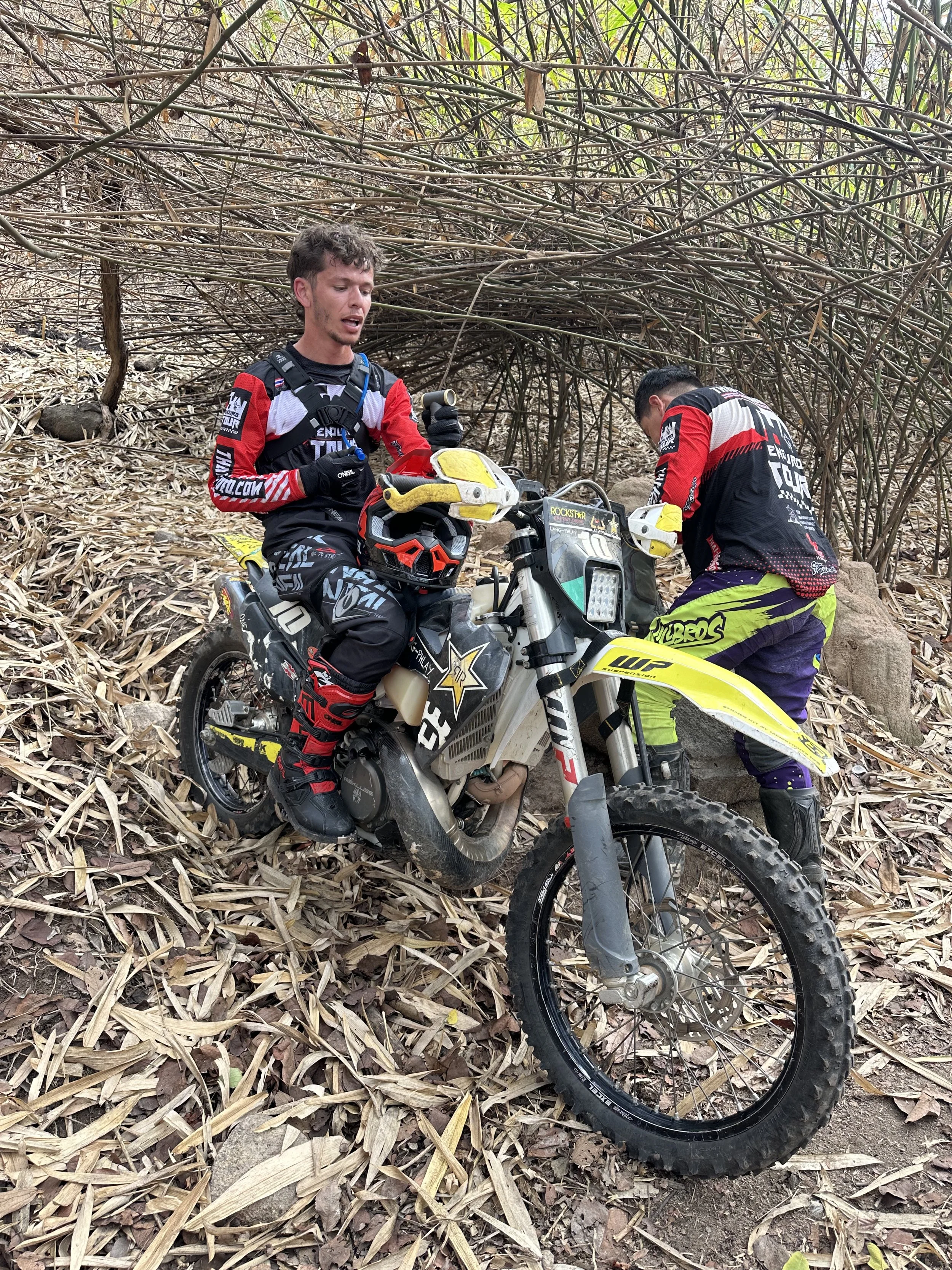 Two motocross riders, dressed in colorful gear, on a dirt trail surrounded by dry leaves and branches, one sitting on his yellow and black dirt bike and the other standing next to the bike, both in a wooded area.