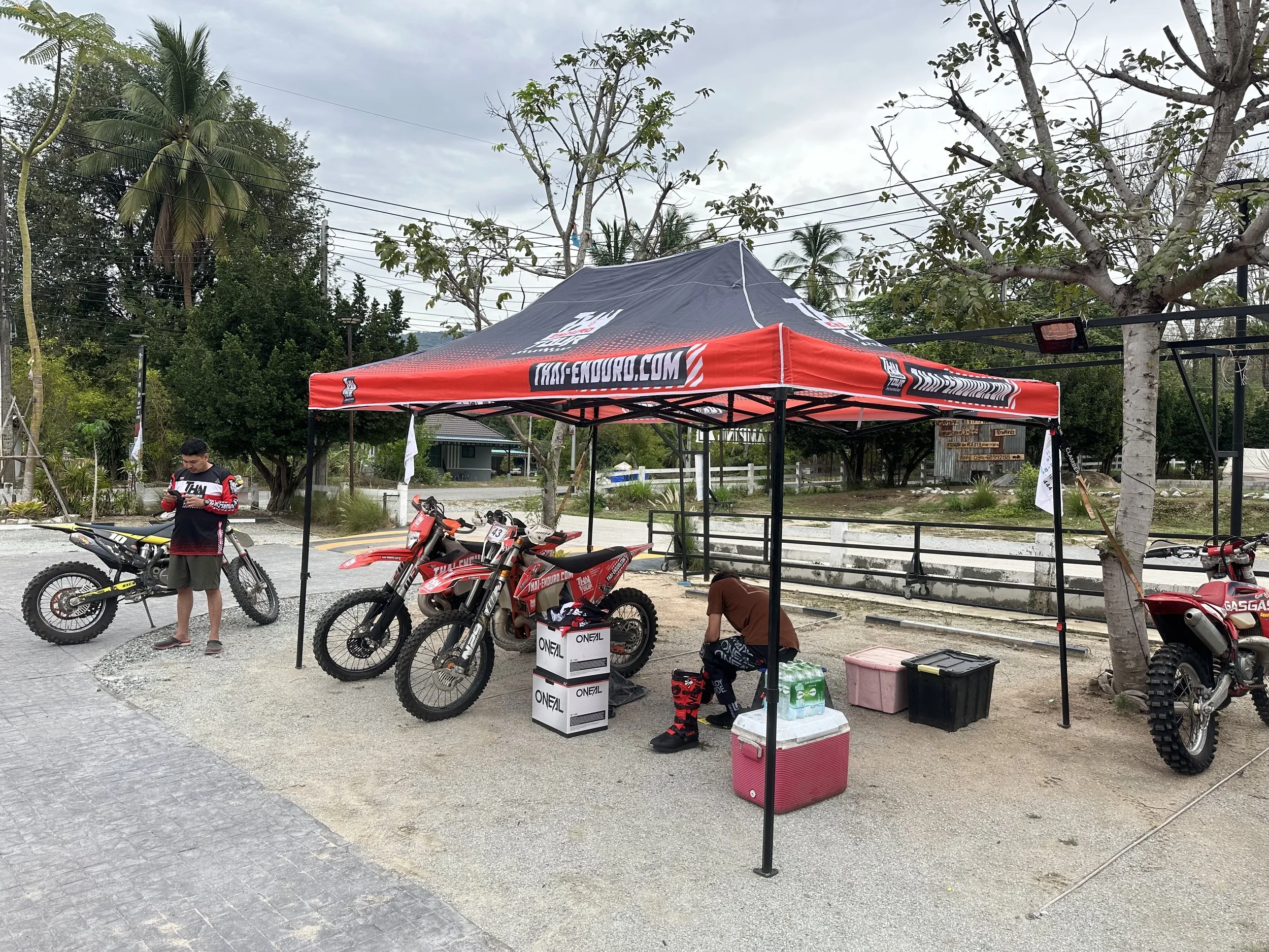 A red and black Honda Enduro tent set up on a dirt area with motocross bikes and a person sitting under the tent. Another person standing nearby on a motorcycle, with trees and a cloudy sky in the background.