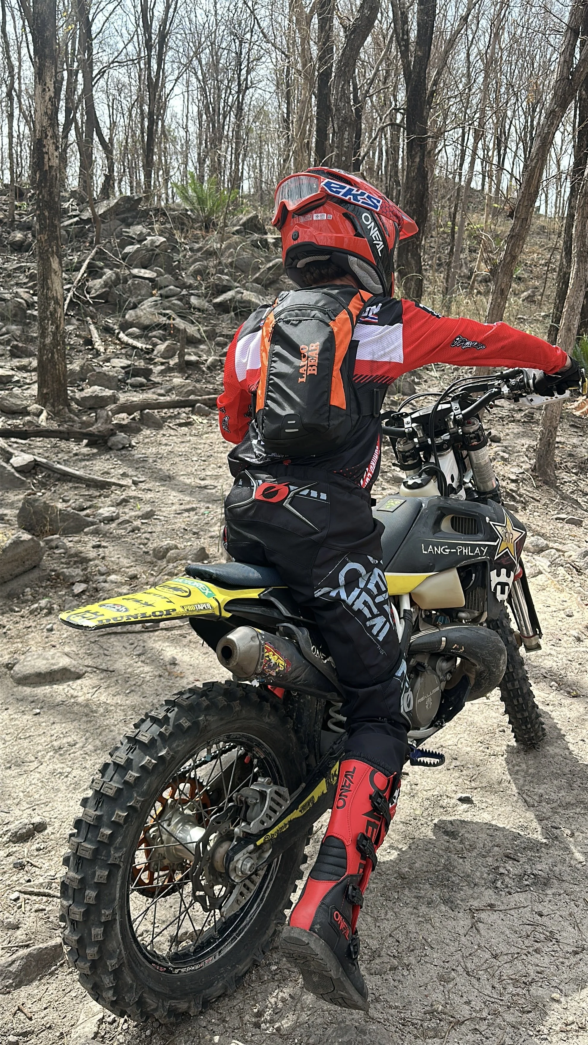 A person in full motocross gear, including a red helmet, red and black riding suit, and red riding boots, sitting on a dirt bike in a wooded area with a rocky trail.