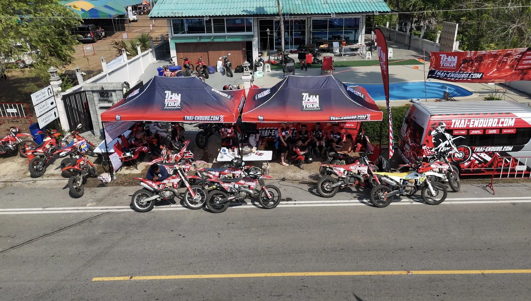 A motorcycle team setup with red canopies displaying 'THAI ENDURANCE' branding, motorcycles parked in front, and team members working or resting around the area. There is a swimming pool, vehicles, and a building in the background.