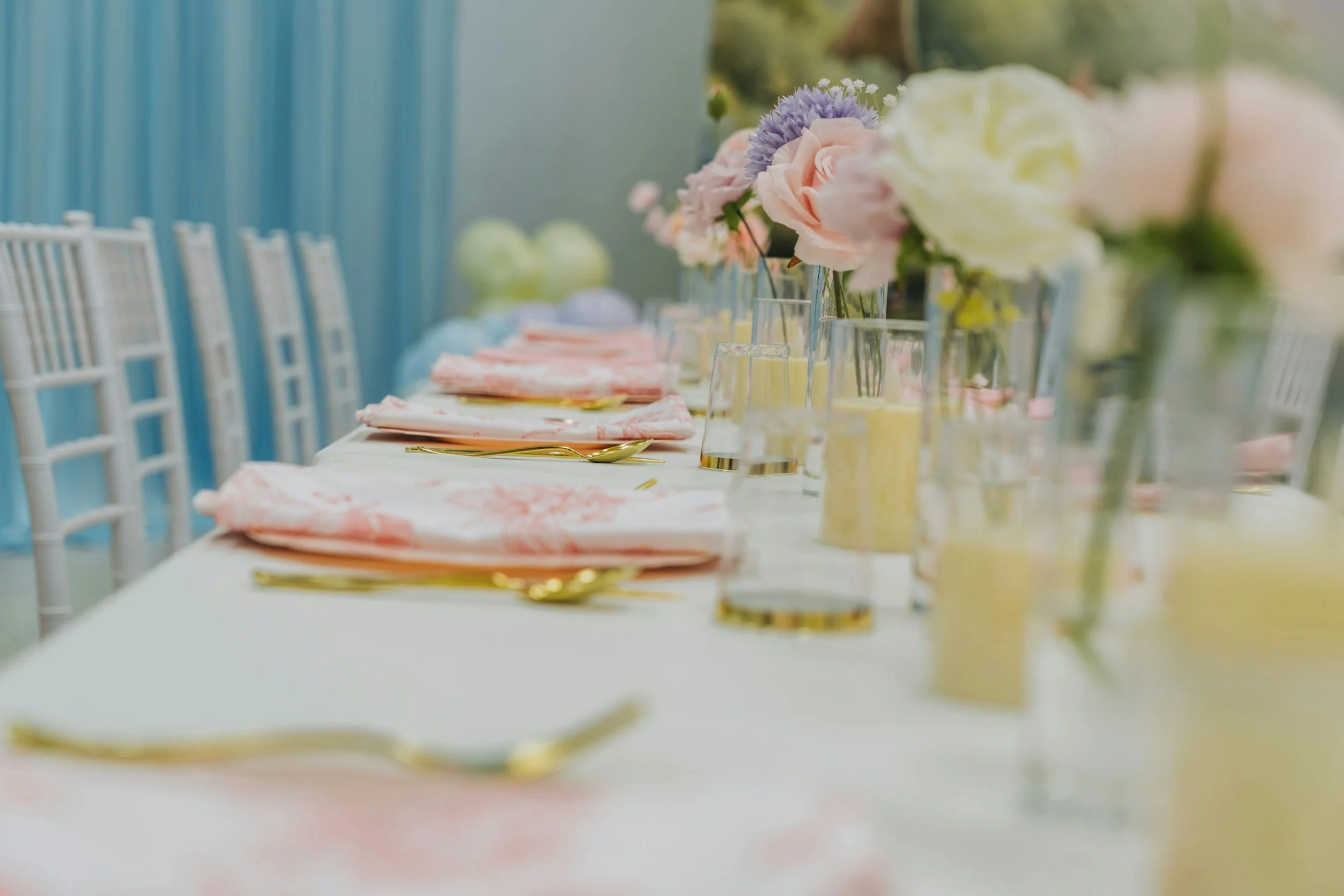 A long table set for a celebration with pink, purple, and white flowers in vases, gold flatware, and pastel napkins, with white chairs and a light blue backdrop.
