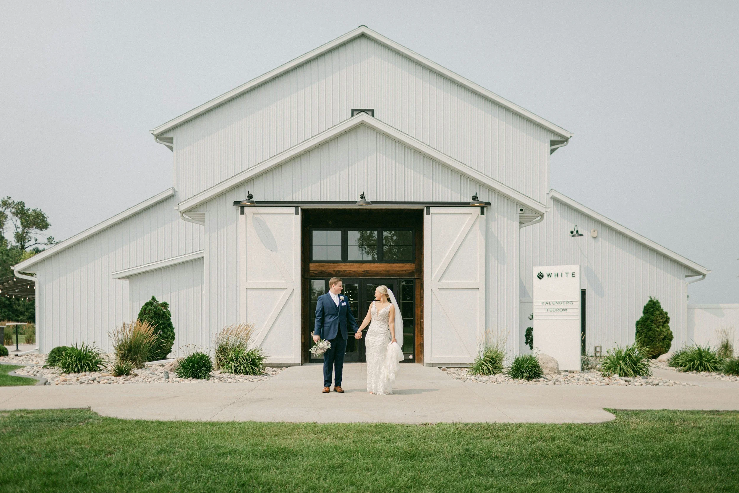 A bride and groom holding hands and looking at each other in front of a white barn with sliding doors at a wedding venue.
