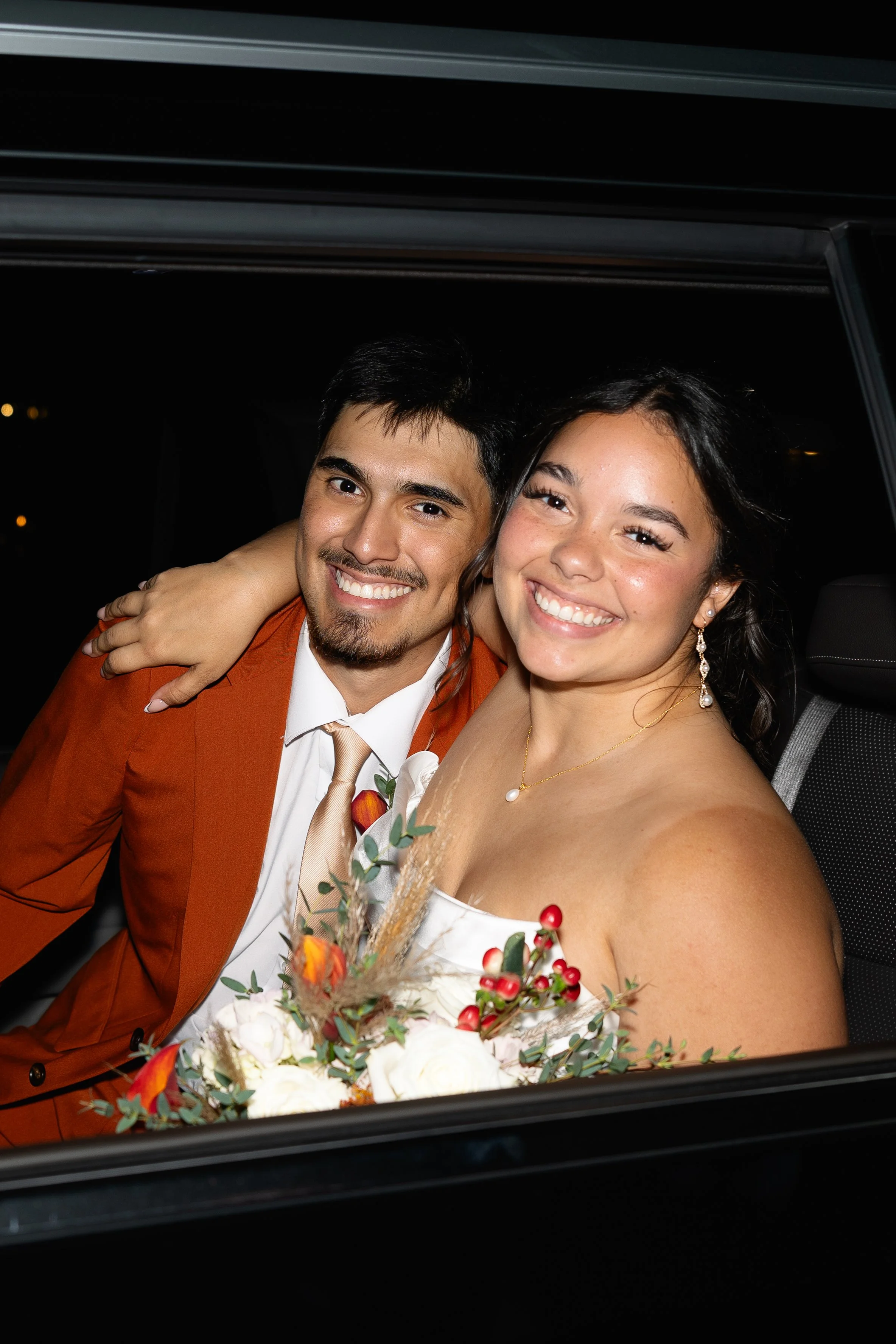 Smiling bride and groom sitting in a vehicle at night, dressed in formal attire, with the woman holding a bouquet of flowers. Blush Your Heart Wedding Company Katy, Texas